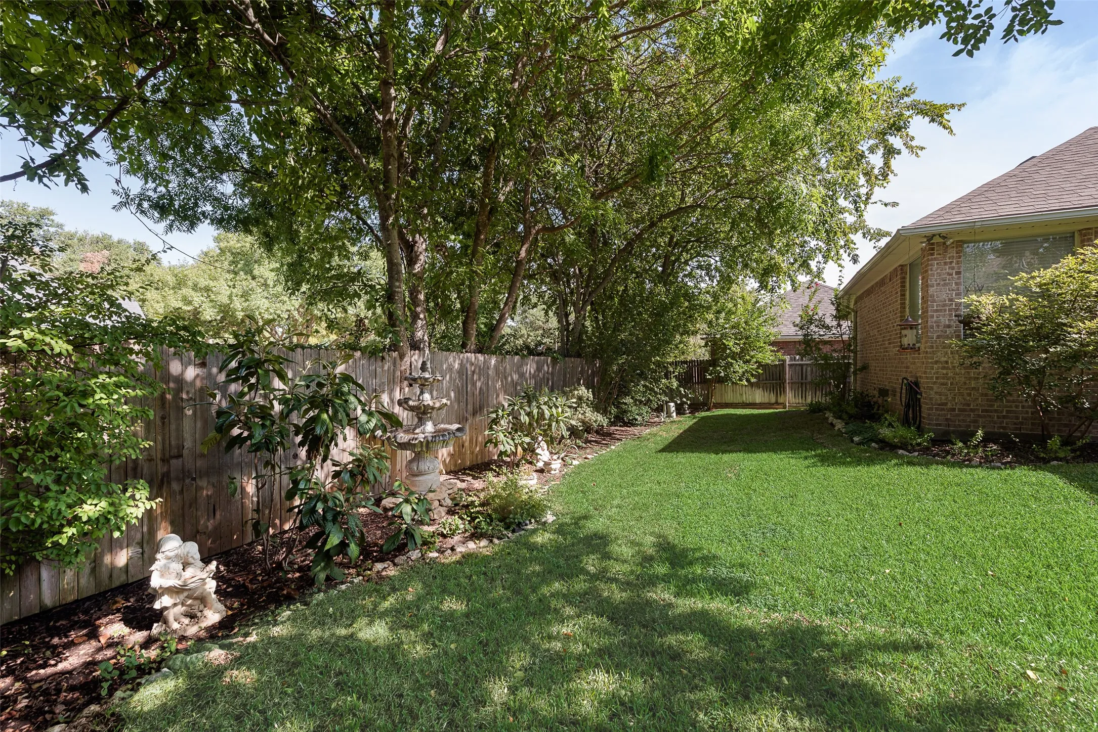 Backyard featuring a manicured lawn, established shade trees, wood privacy fencing, and a tiered decorative fountain
