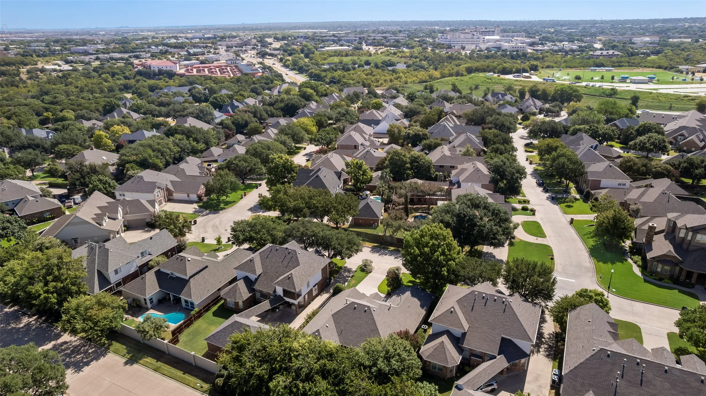 Aerial perspective of a residential neighborhood featuring multi-story dwellings, paved roadways, mature tree canopy, and private swimming pools
