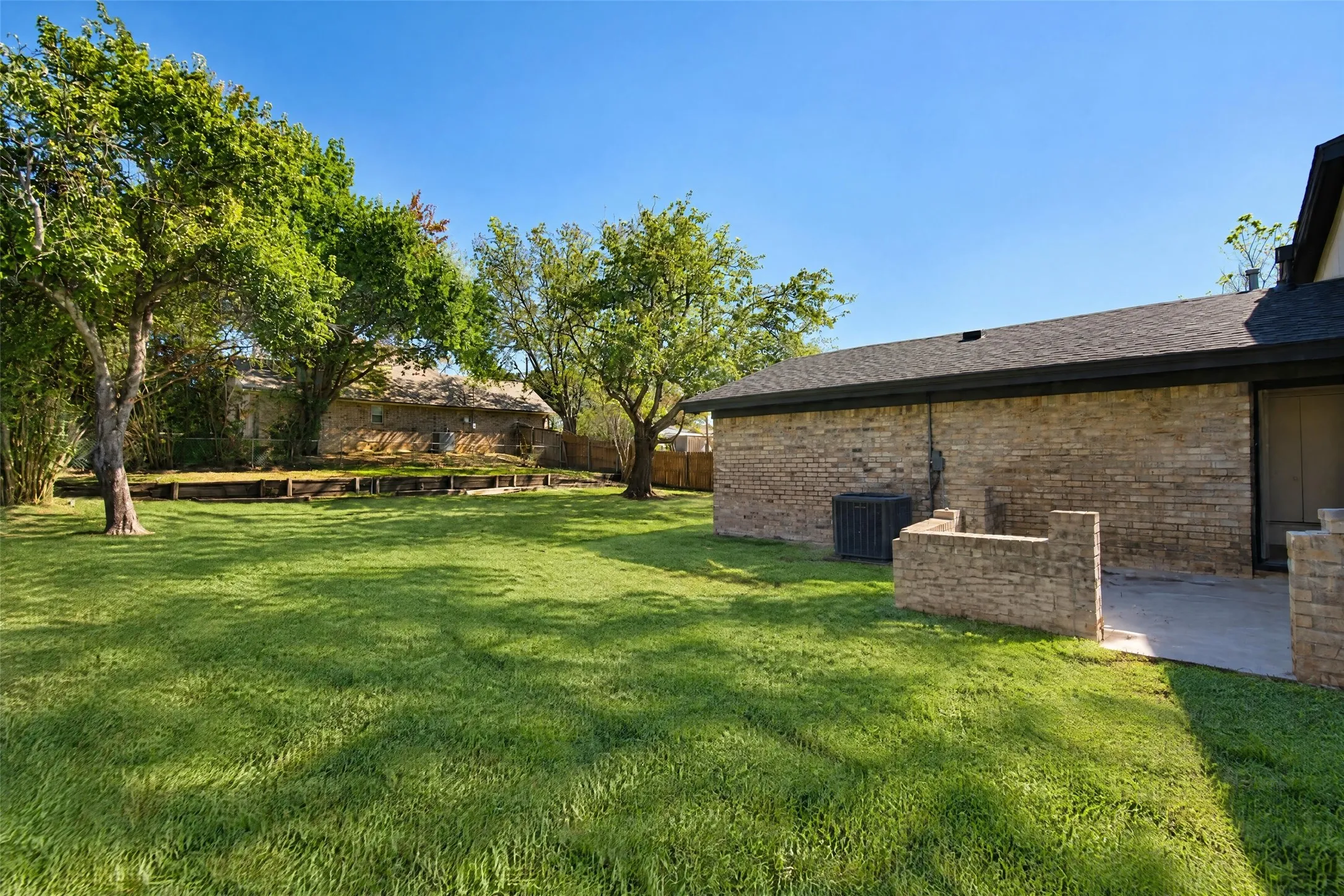 Expansive backyard featuring a brick exterior, stone-walled patio, timber retaining wall, and mature trees