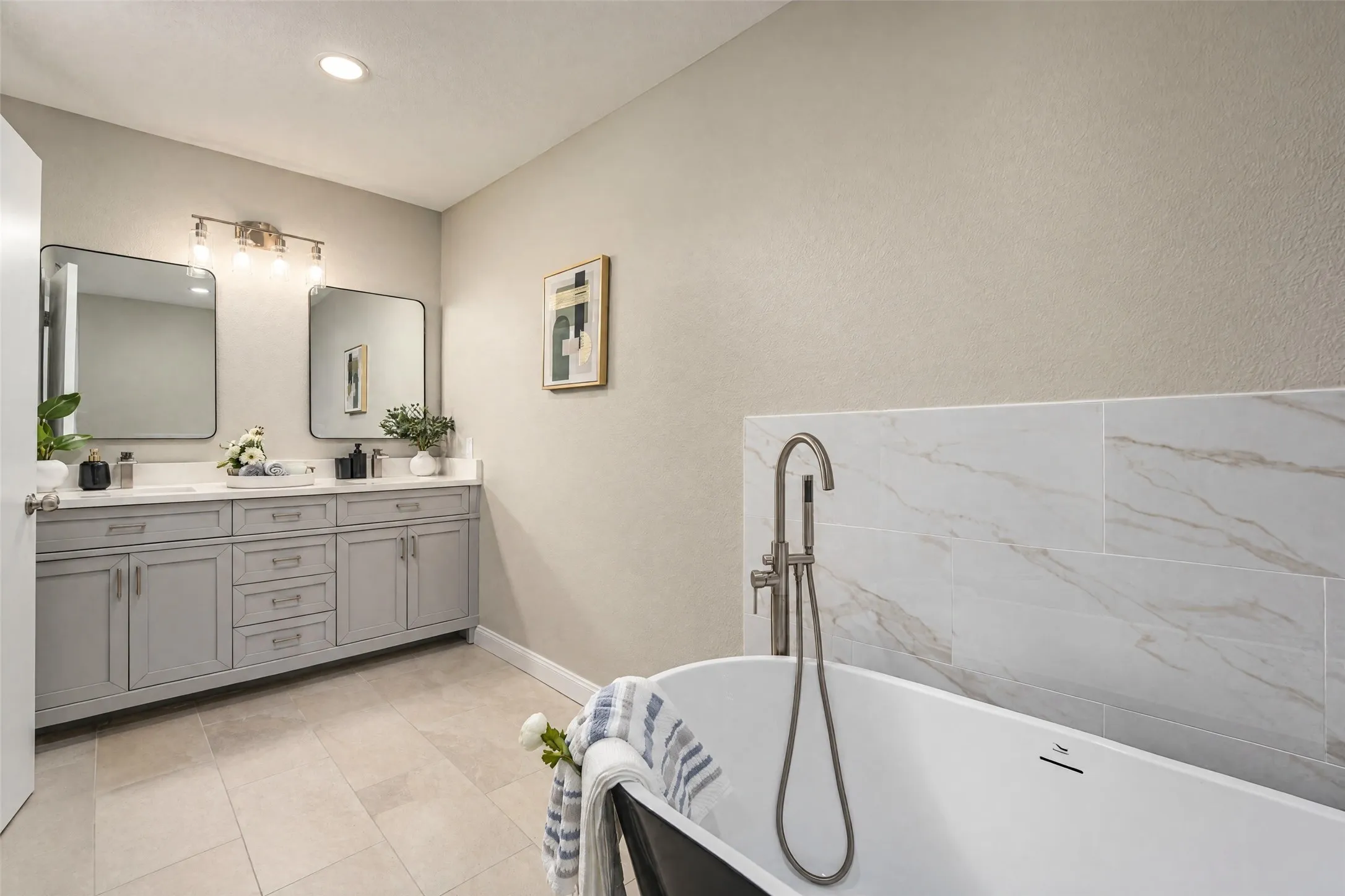 Freestanding oval bathtub with a floor-mounted brushed nickel faucet, light-toned large-format tile flooring, double-sink vanity with grey cabinetry, rectangular mirrors, and a tiled tub surround
