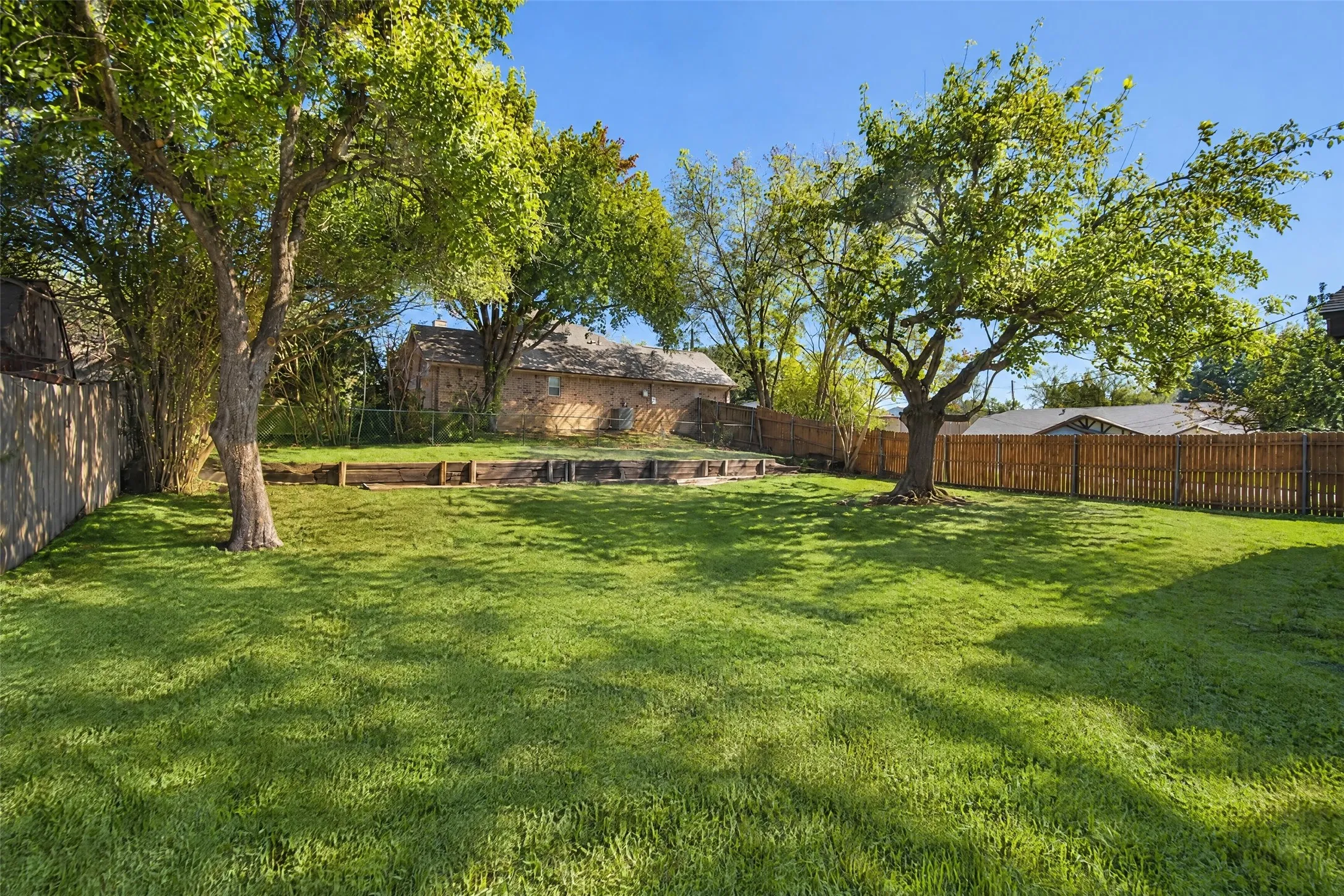 Expansive backyard featuring a large grass lawn, mature trees, wood privacy fencing, and tiered timber retaining walls