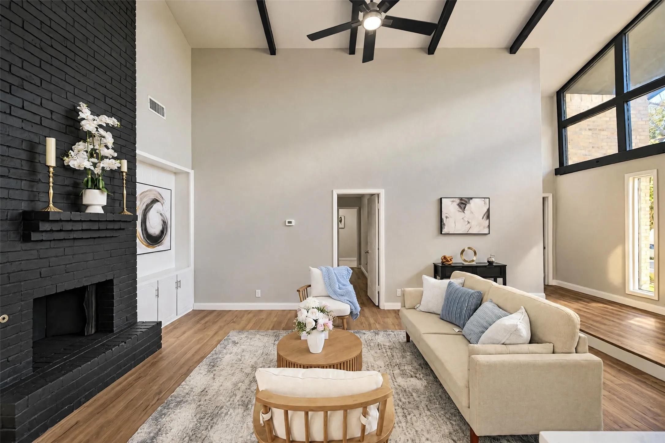 Vaulted ceiling with exposed beams, floor-to-ceiling painted brick fireplace, built-in cabinetry, wood-finish flooring, and expansive multi-pane windows