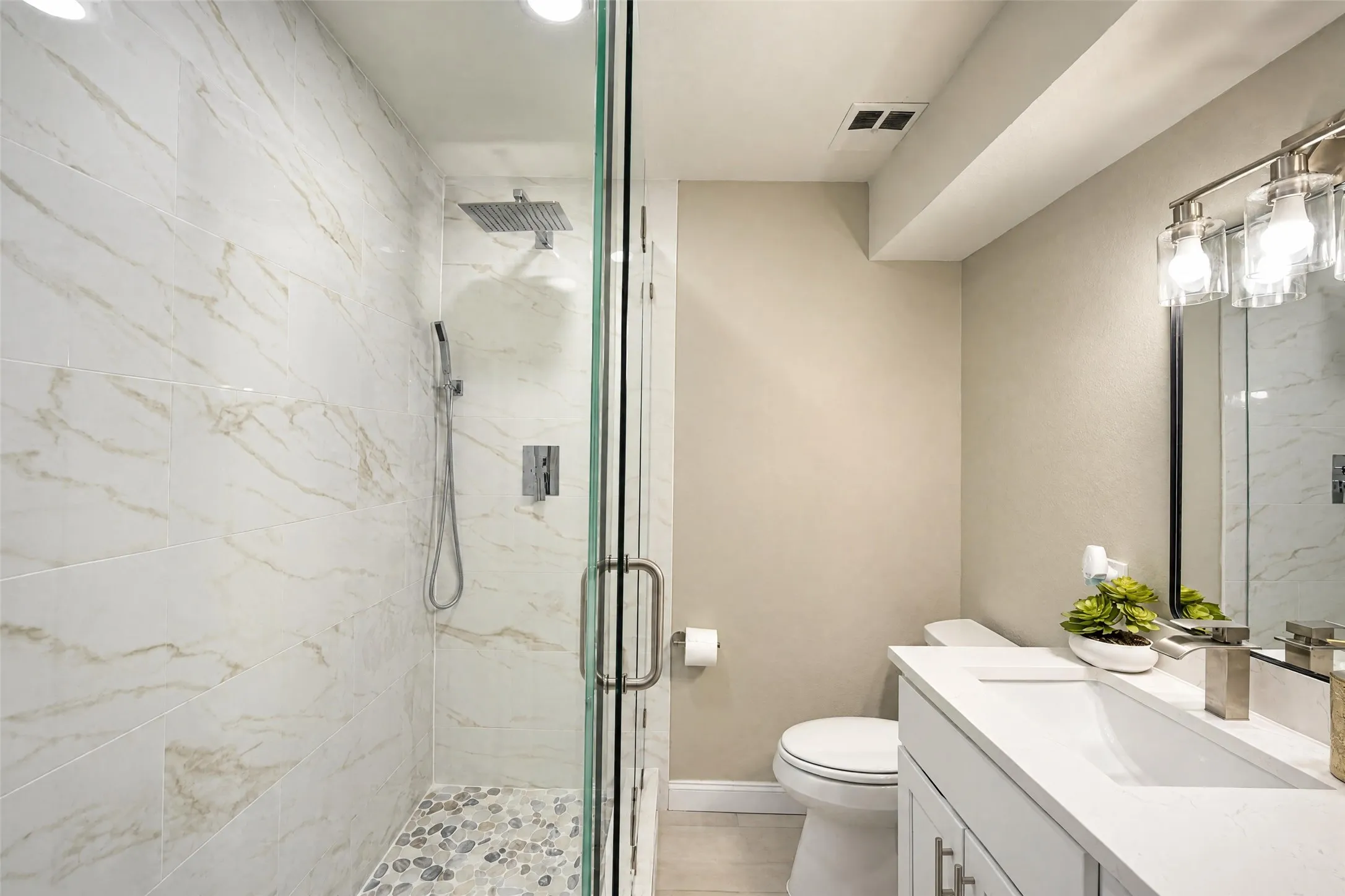 Walk-in shower featuring glass enclosure, rain-style showerhead, and stone-tiled floor, vanity with stone-composite countertop, and wall-mounted multi-light fixture