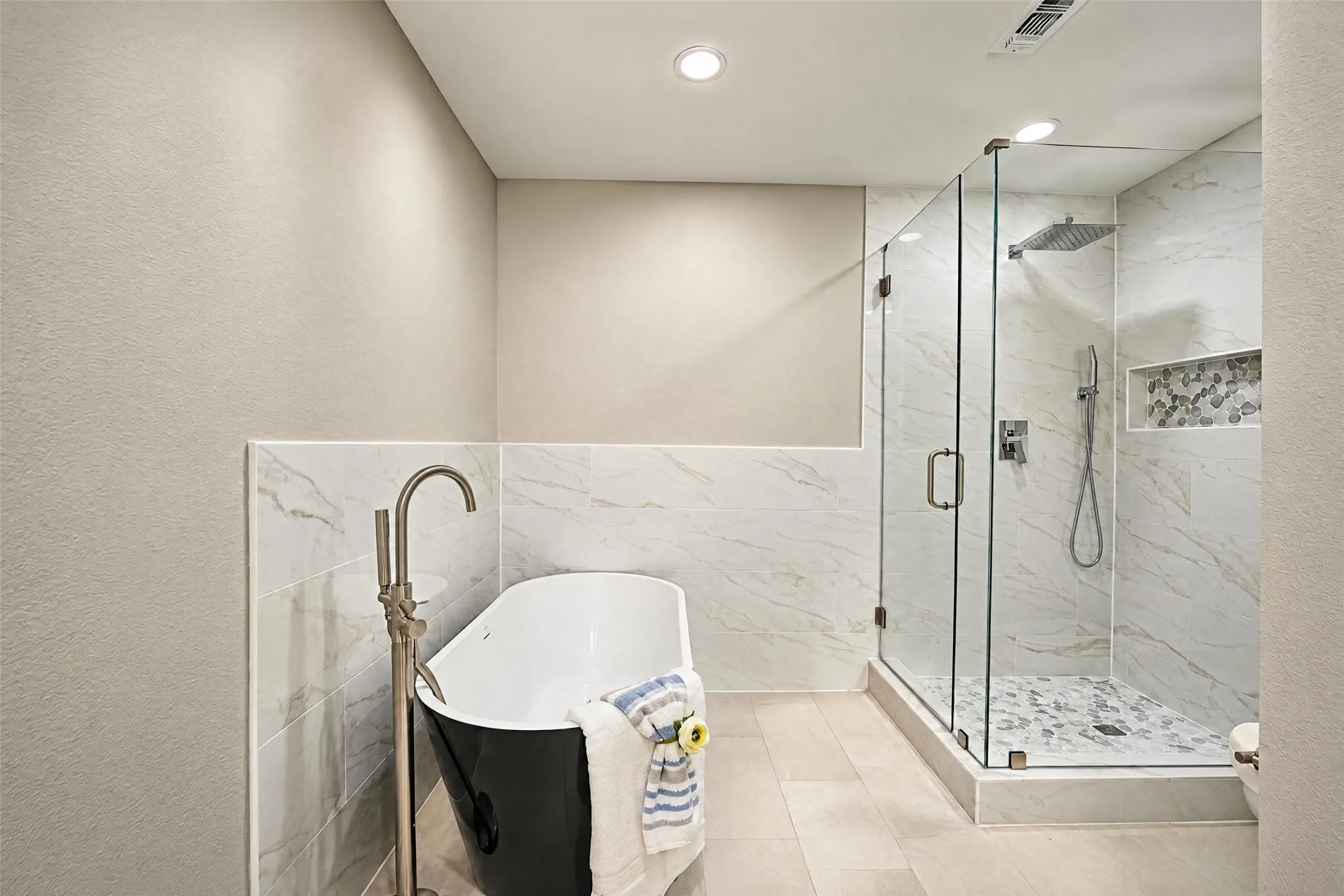 Freestanding soaking tub with floor-mounted faucet, walk-in glass-enclosed shower, wall-mounted rainfall showerhead, stone-finish tile surround, and built-in shower niche