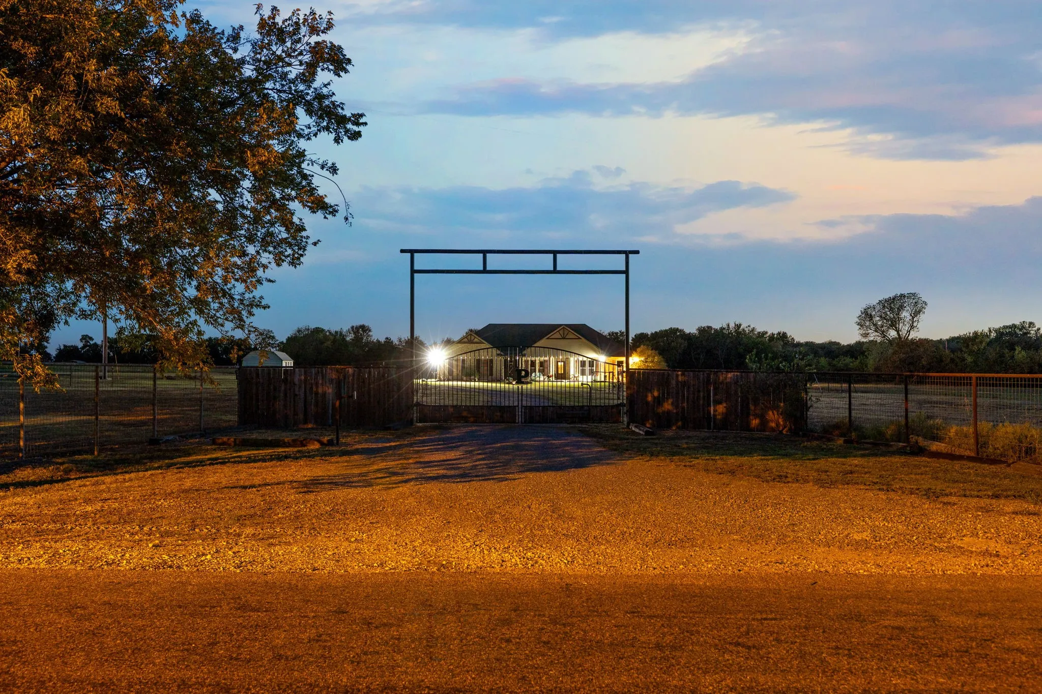 Gated entrance featuring a metal overhead arch, extensive perimeter fencing, and a gravel drive