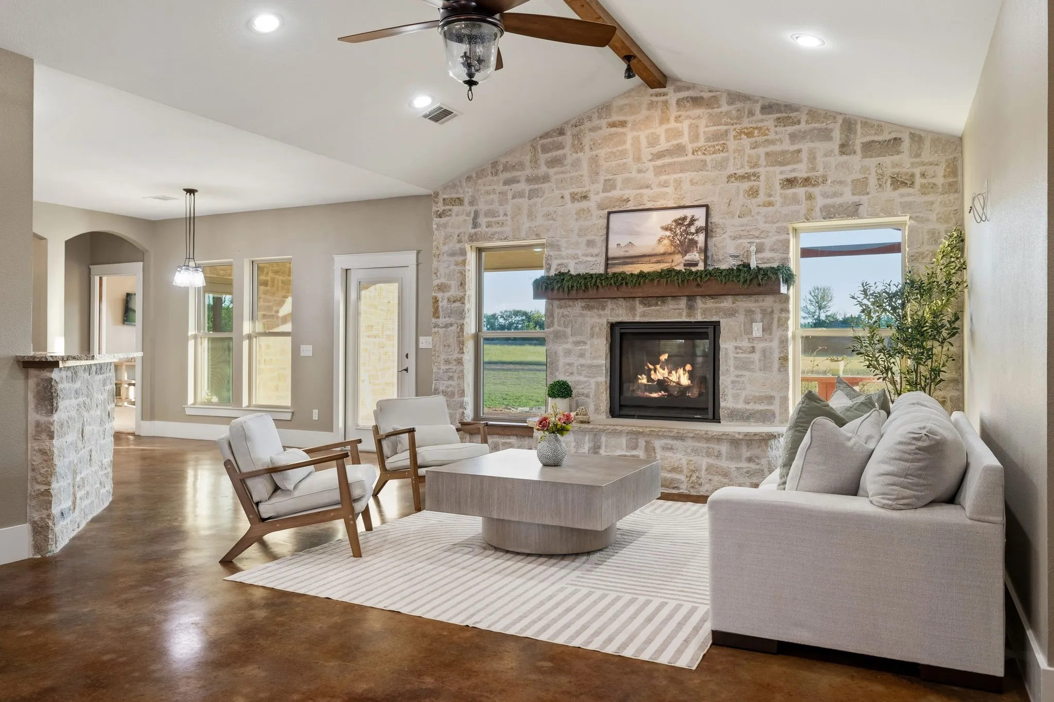 Vaulted ceiling with exposed wood beam, floor-to-ceiling stone fireplace with timber mantel, polished concrete flooring, and recessed lighting