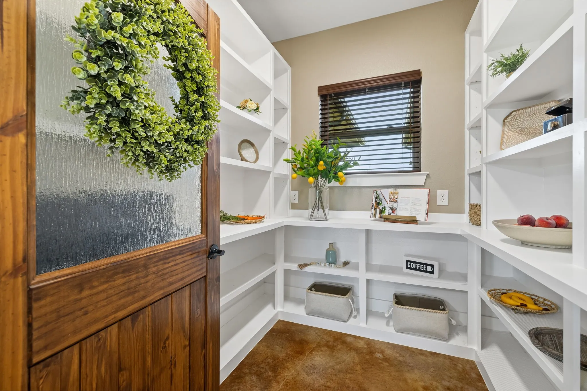 Pantry featuring floor-to-ceiling white custom built-in shelving, concrete flooring, and a natural wood-finish door with textured glass paneling