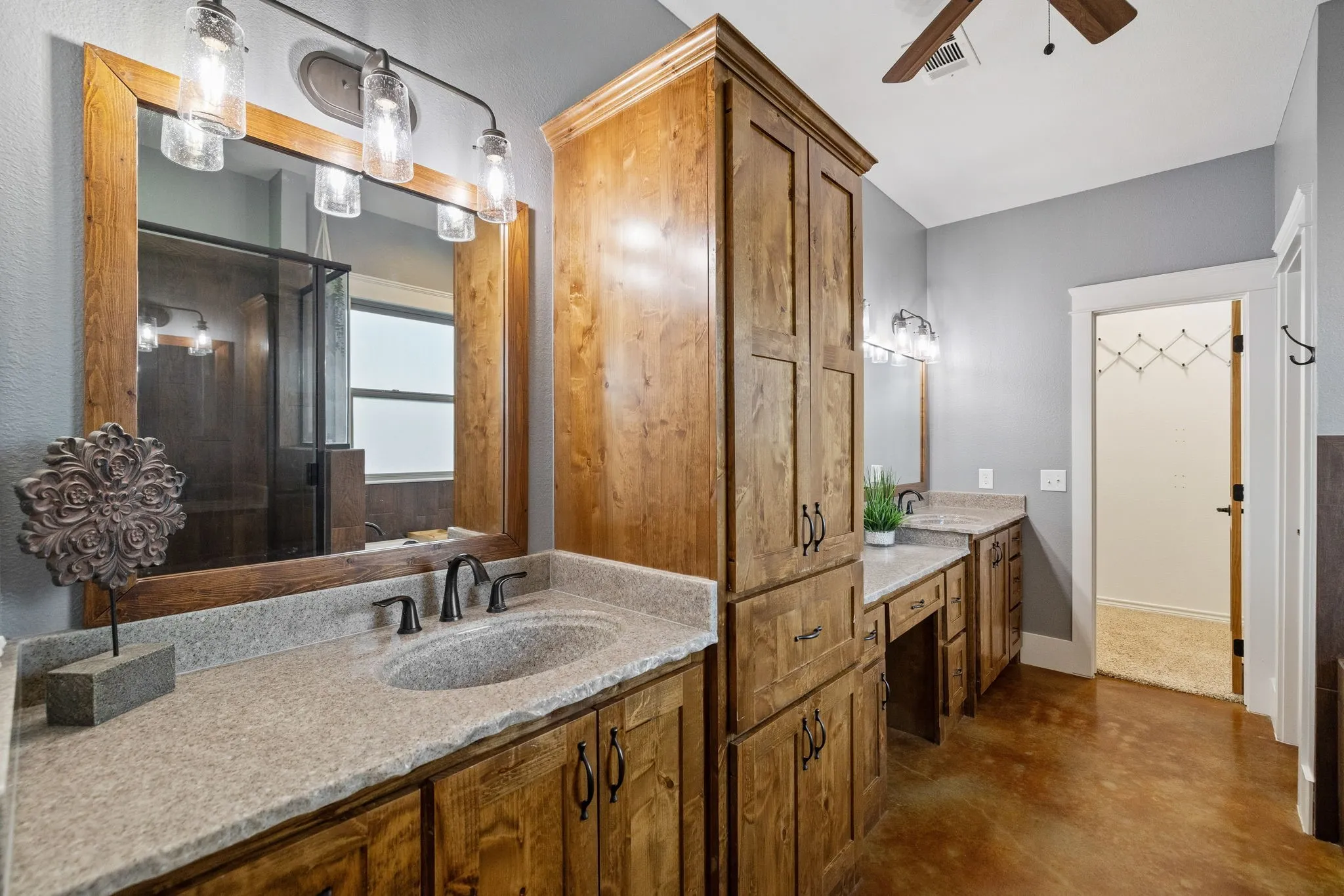 Custom wood cabinetry with integrated tall storage, stone-finish countertops, dual mirrors with wood frames, multi-light vanity fixtures, and stained concrete flooring