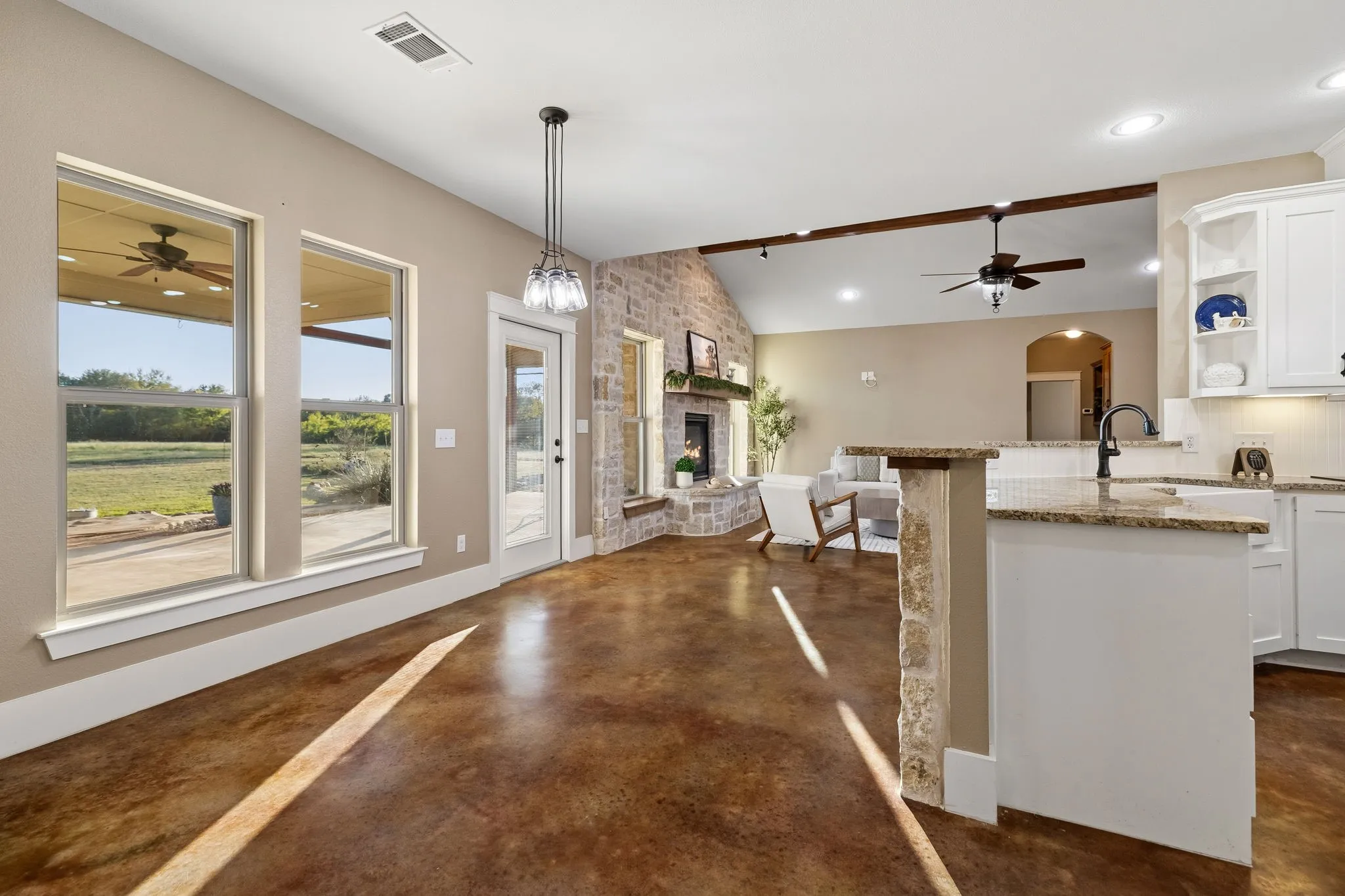 Open-concept living space featuring stained concrete flooring, stone-faced fireplace, kitchen island with stone countertop, and multi-pane windows