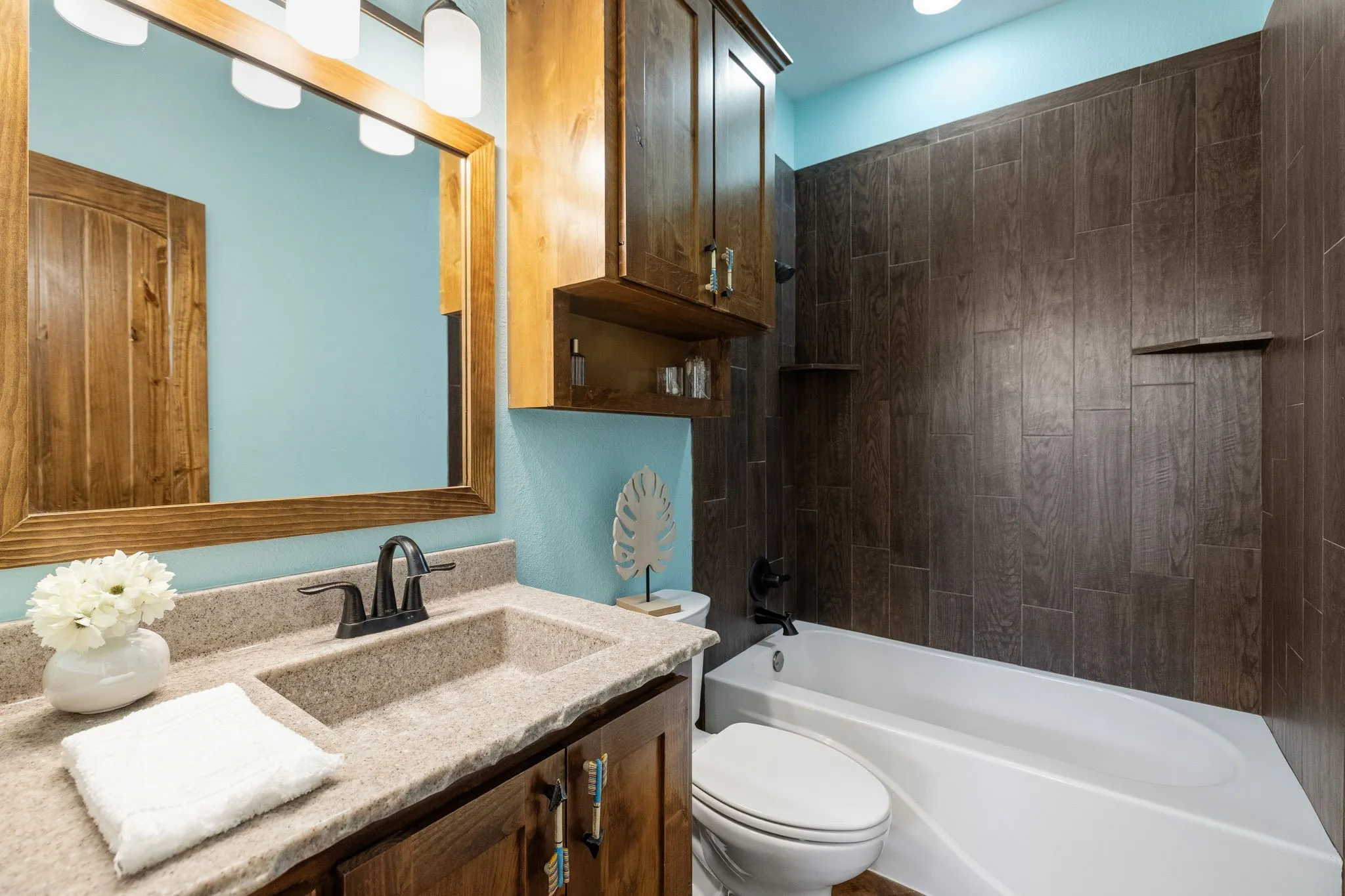 Bathroom featuring a stone-finish countertop with an integrated sink, wood-finish cabinetry, a large wood-framed mirror, and a shower-tub combination with dark tile surround