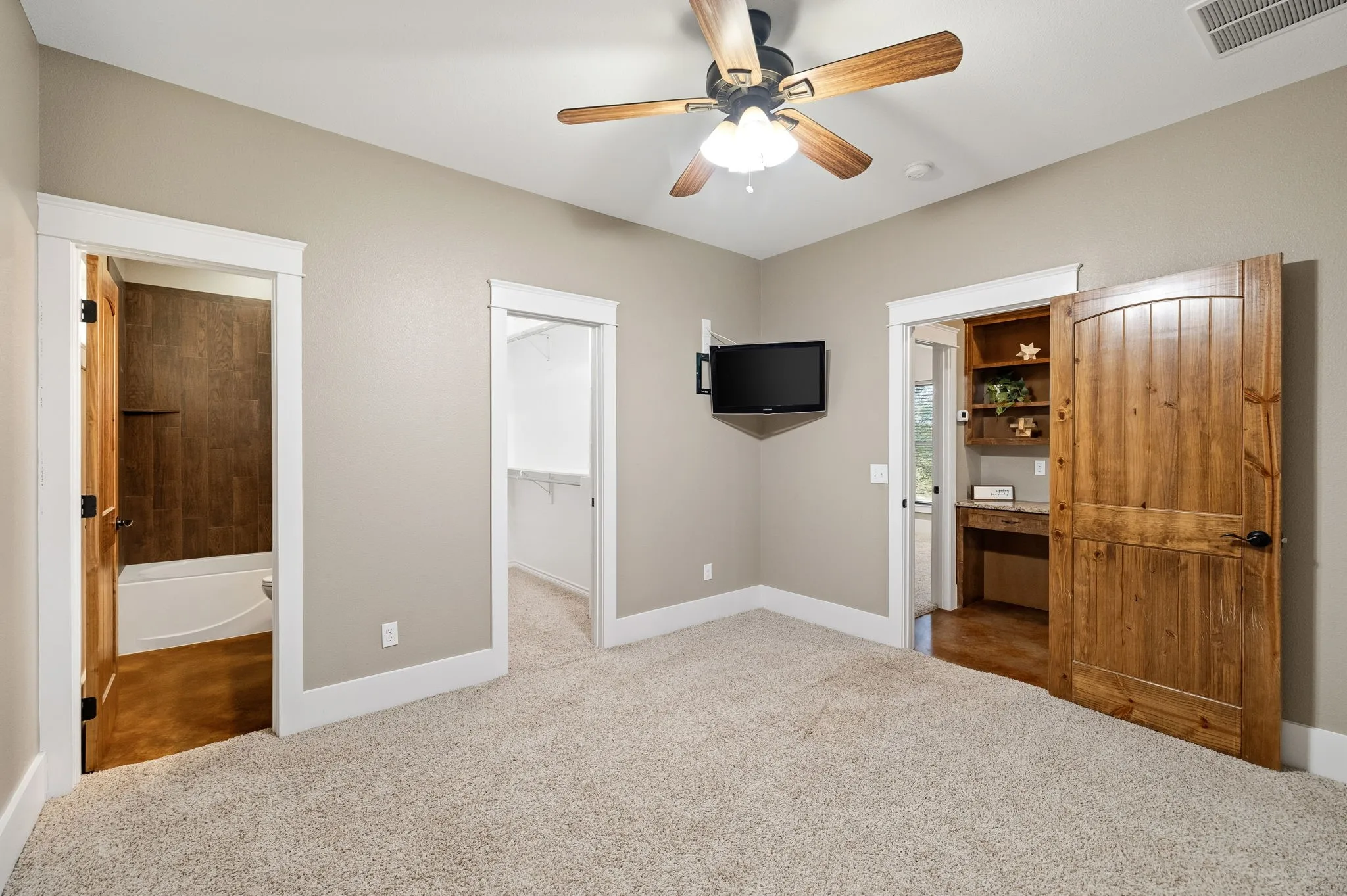 Neutral wall tones complemented by wood-finish interior doors, ceiling fan with integrated lighting, wall-to-wall carpeting, and a built-in wooden desk with shelving