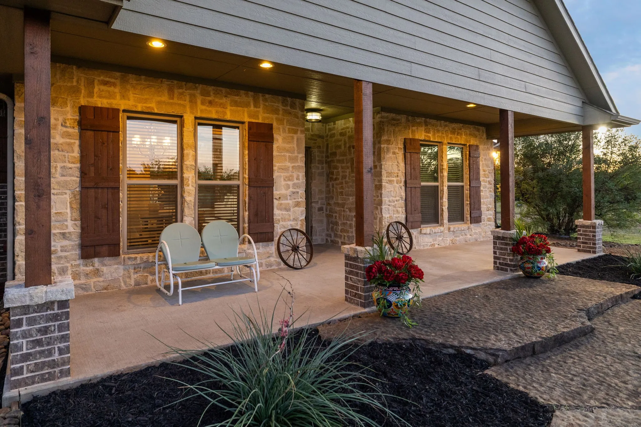 Covered front porch featuring natural stone facade, wood-finish support posts, brick masonry column bases, and decorative window shutters