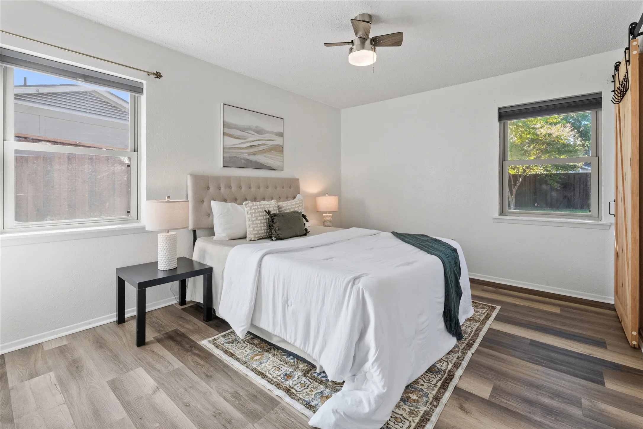Bedroom with wood-finish flooring, dual windows with roller shades, a ceiling fan with integrated lighting, and a sliding barn-style door