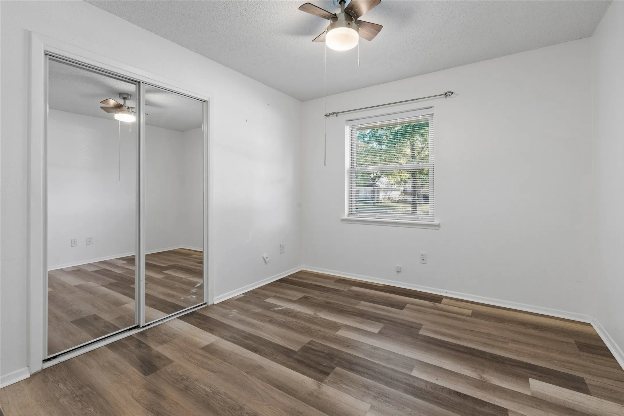 Room featuring wood-finish flooring, sliding mirrored closet doors, a single-hung window, and a ceiling fan with integrated light fixture