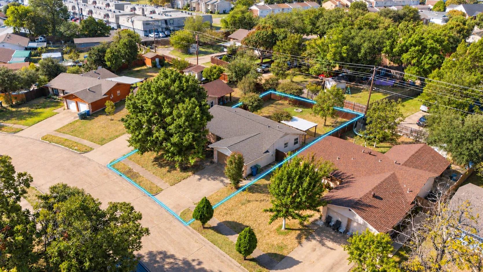Aerial view of a residential property featuring a composition shingle roof, concrete driveway, attached garage, fenced rear yard, and mature tree landscaping