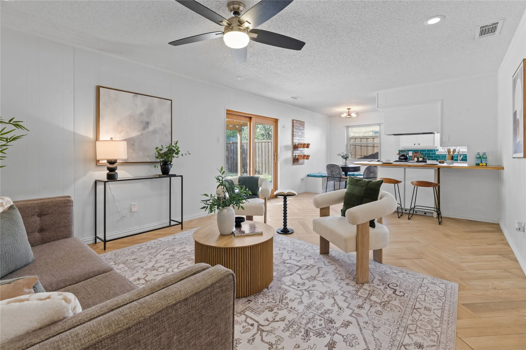 Open-concept interior featuring light wood-finish herringbone flooring, a built-in kitchen breakfast bar, a modern ceiling fan, and sliding glass doors with exterior access