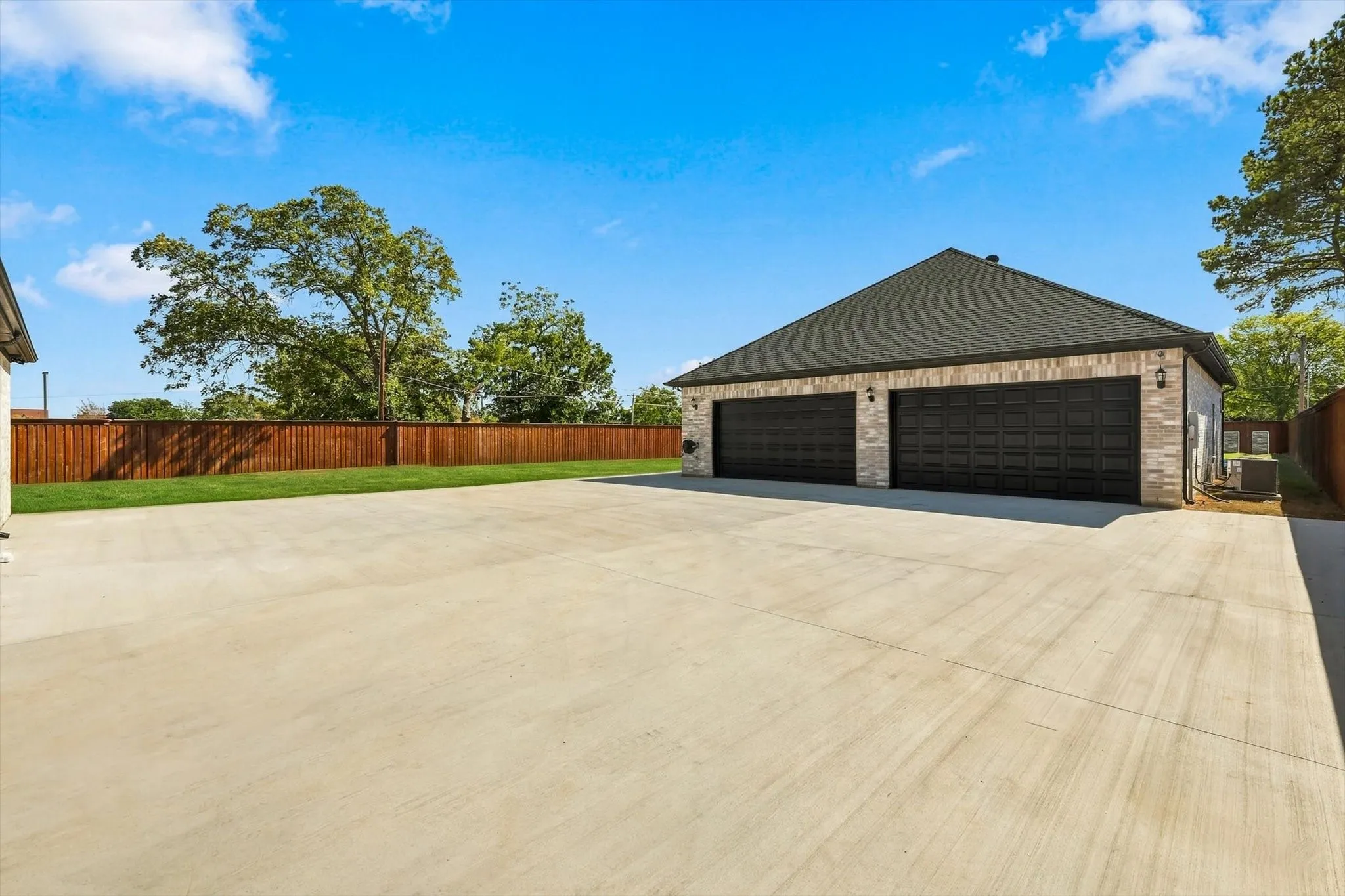 Detached three-car garage with masonry siding, shingle roof, expansive concrete driveway, and wood-paneled privacy fencing