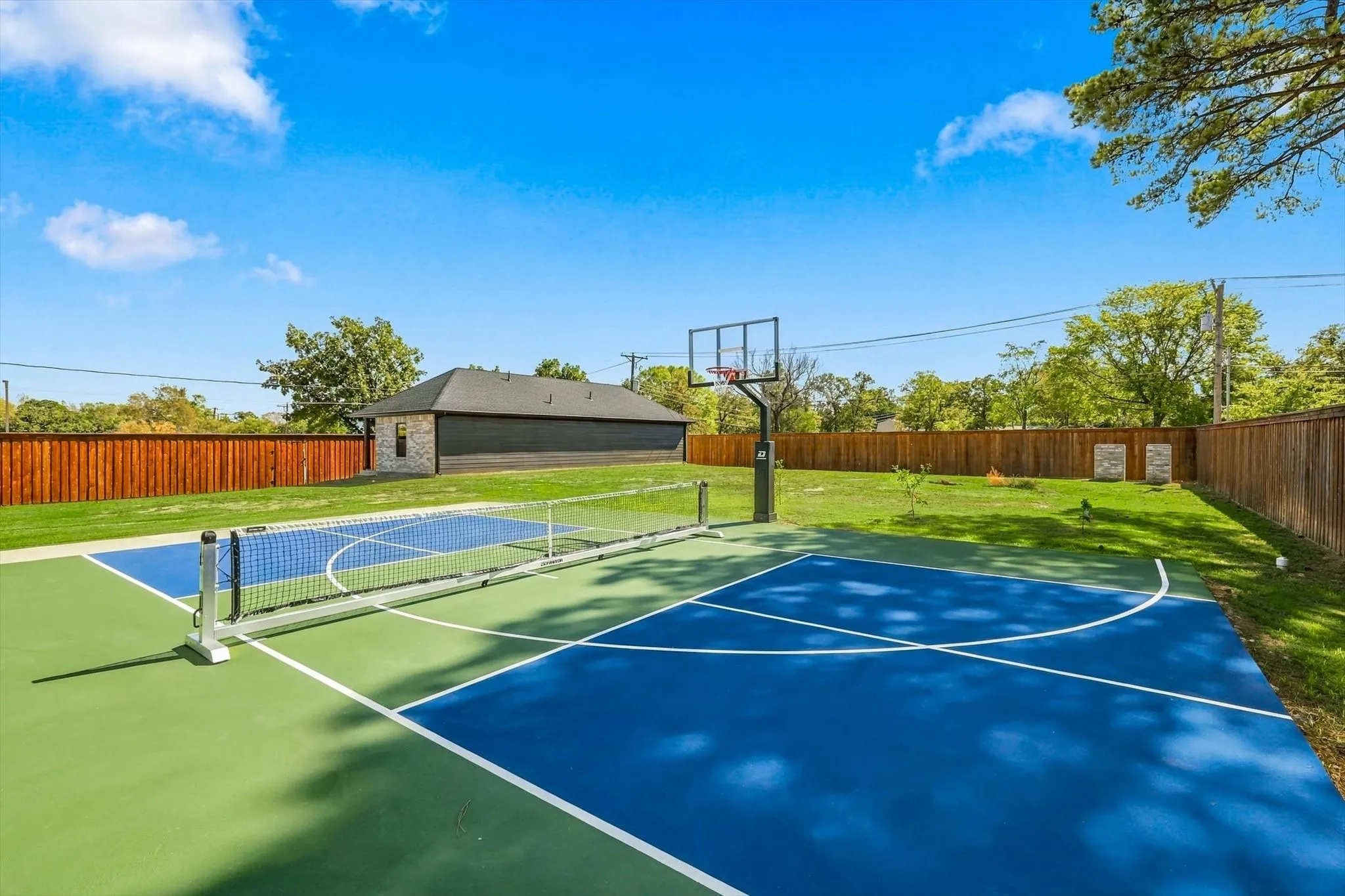 Multipurpose outdoor sports court featuring a pickleball net and built-in basketball hoop, set within a fenced backyard with a detached garage