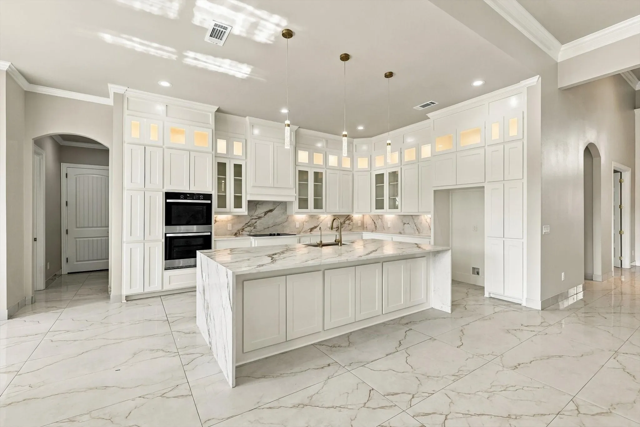 Kitchen featuring white cabinetry with glass-front uppers, waterfall-edge island, stone-finish countertops and backsplash, tile flooring, and integrated double wall ovens