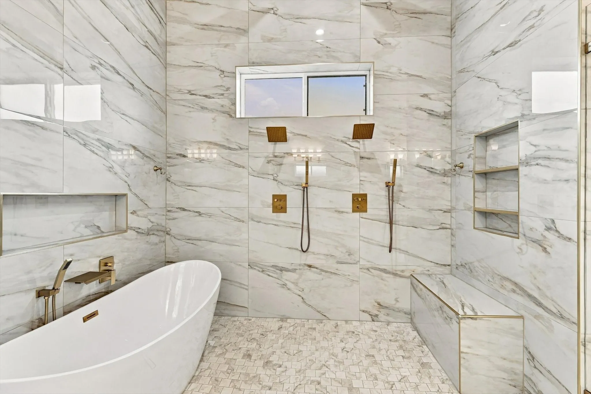 Freestanding soaking tub with wall-mounted metallic fixtures, floor-to-ceiling large-format tile surround, dual rainfall showerheads with handheld sprayers, built-in tiled bench, and recessed wall shelving