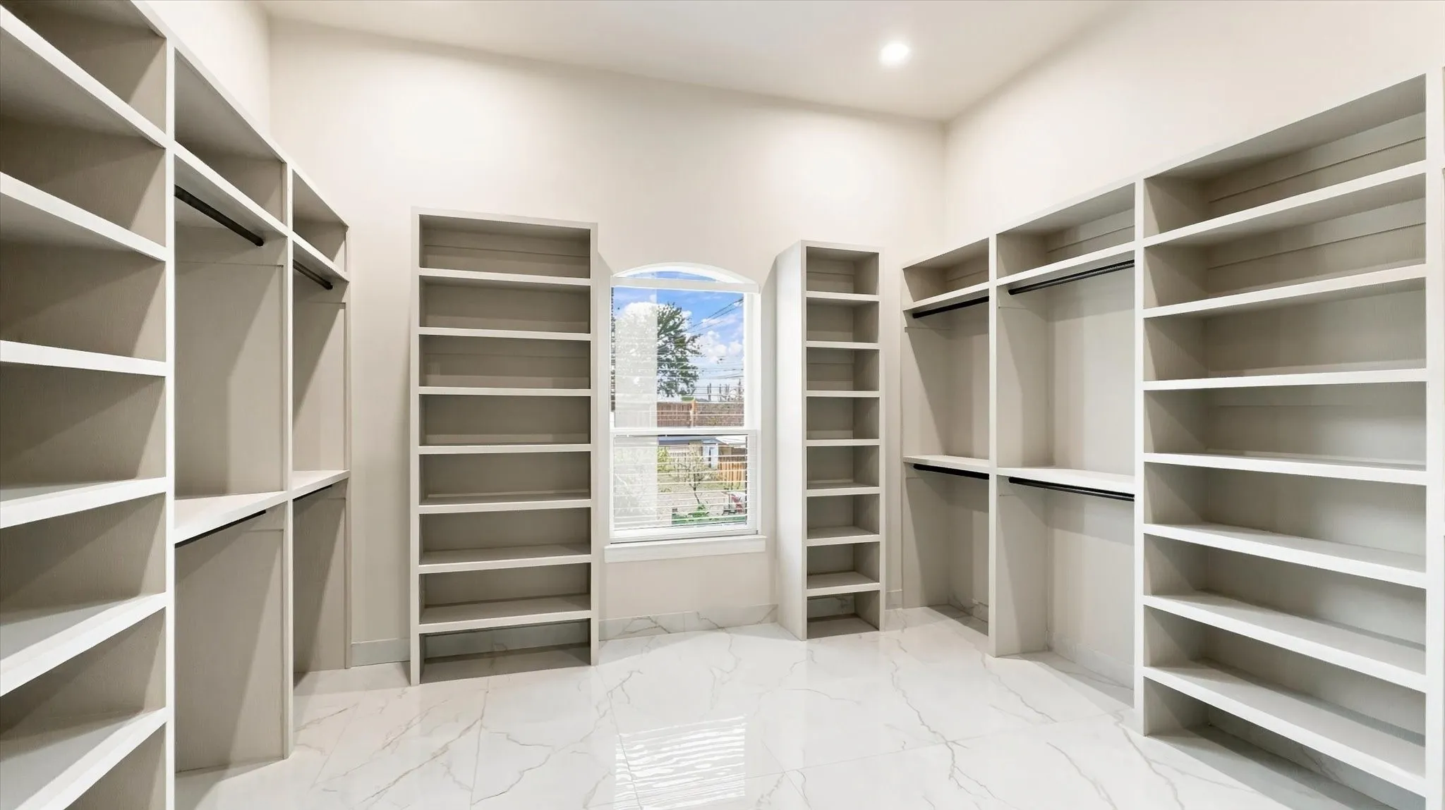 Custom storage room featuring extensive built-in cabinetry, floor-to-ceiling shelving, hanging rods, an arched window, and large-format tile flooring