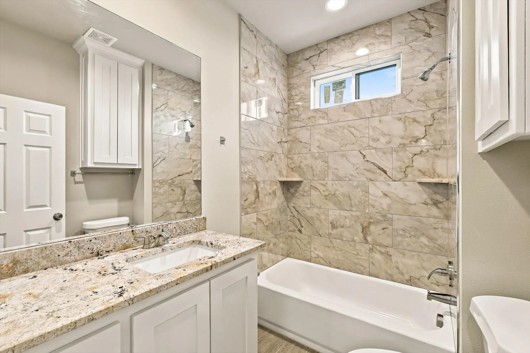 Bathroom featuring a stone-look countertop with an integrated basin, white shaker-style cabinetry, a large wall-mounted mirror, and a shower-tub combination finished with full-height stone-look tile and built-in shelving