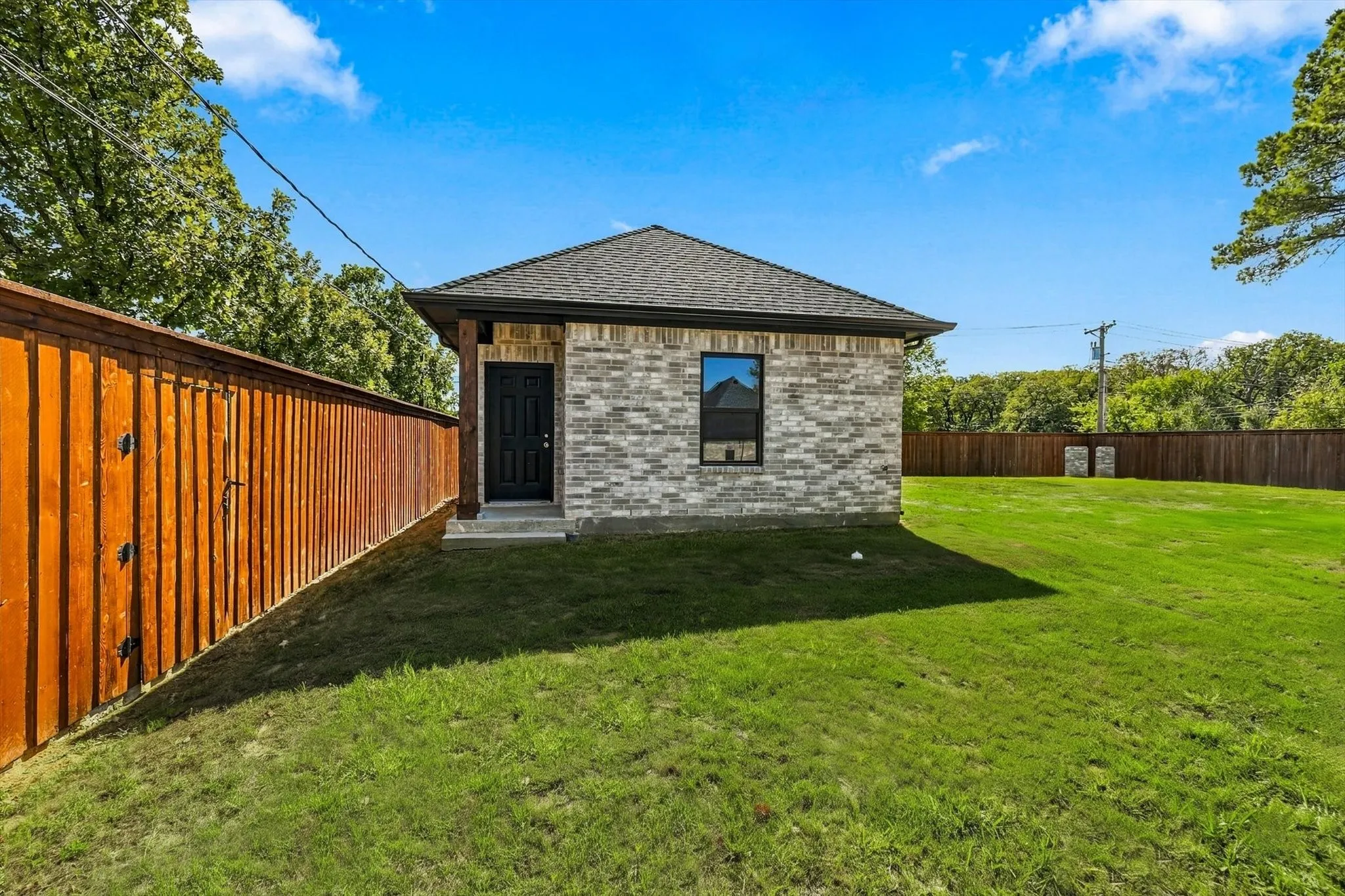 Exterior facade featuring a grey brick finish, a hipped roof, a dark-toned entry door, and a full wood-plank privacy fence surrounding a grass yard