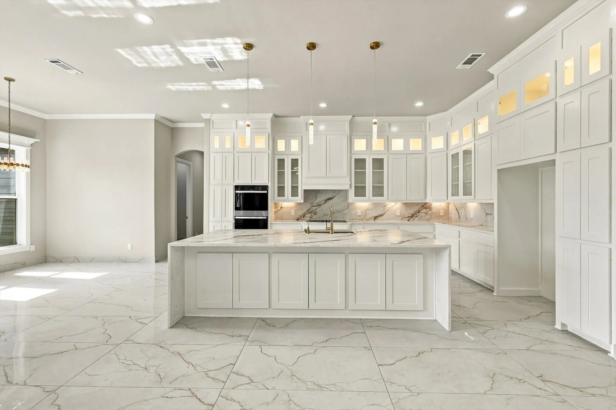 Kitchen featuring white floor-to-ceiling cabinetry, large center island with waterfall edge, integrated wall oven, stone-finish countertops and backsplash, and light-tone tile flooring