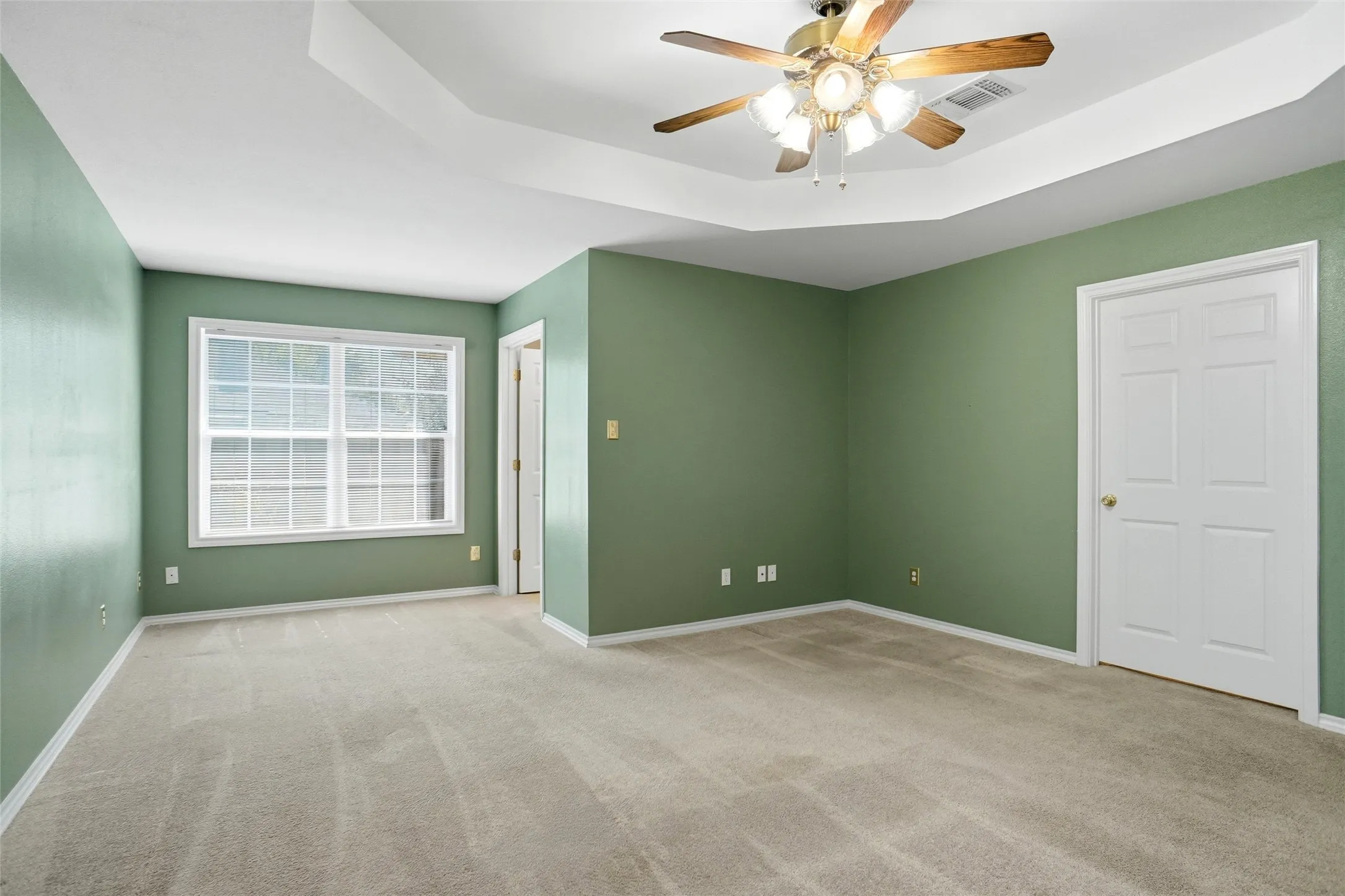 This view of the primary bedroom shows the siitting area.  Tray ceiling with an overhead ceiling fan, neutral carpet flooring, and a multi-pane window