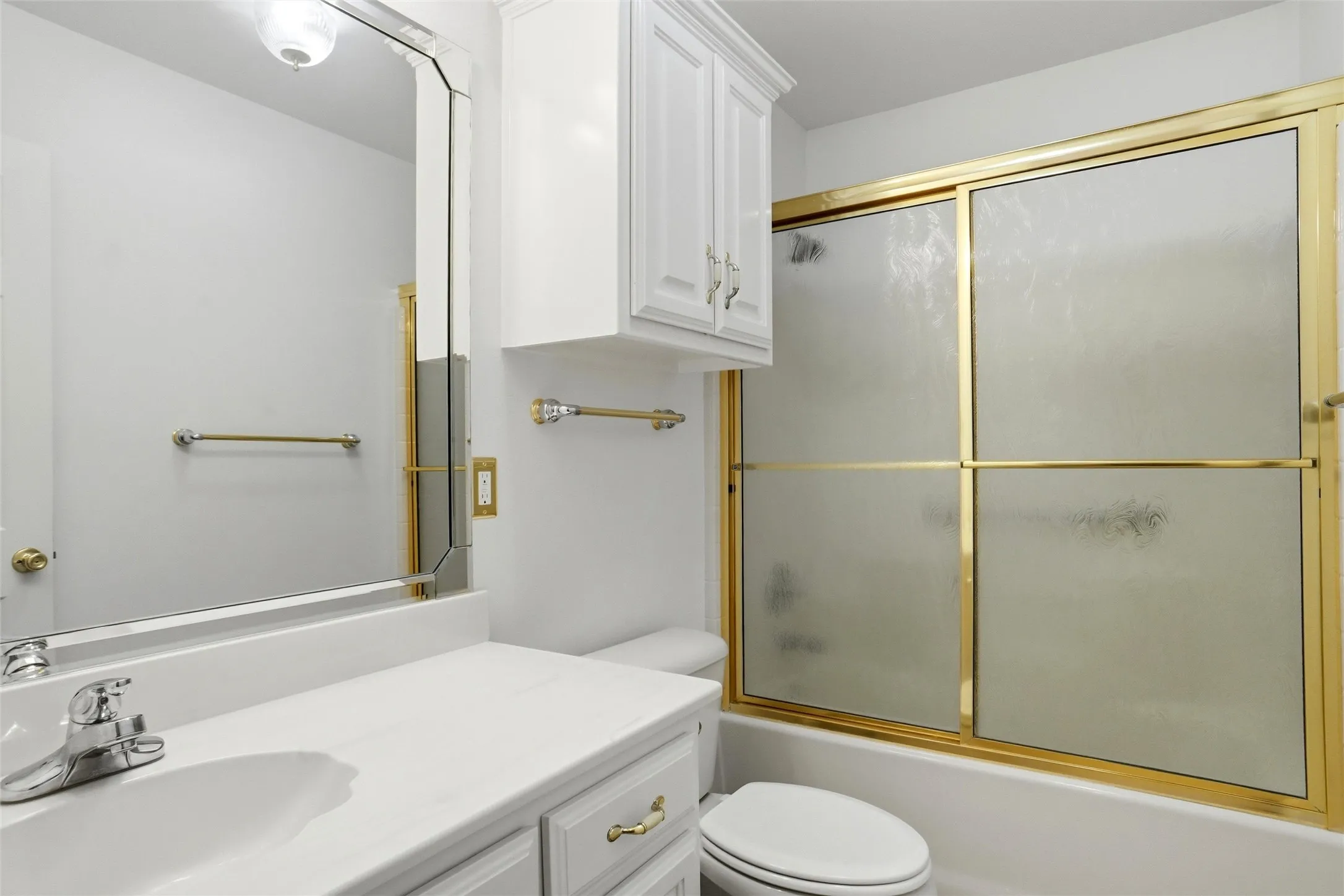 White vanity with an integrated sink and chrome faucet, gold-framed shower with frosted glass doors, and upper cabinetry