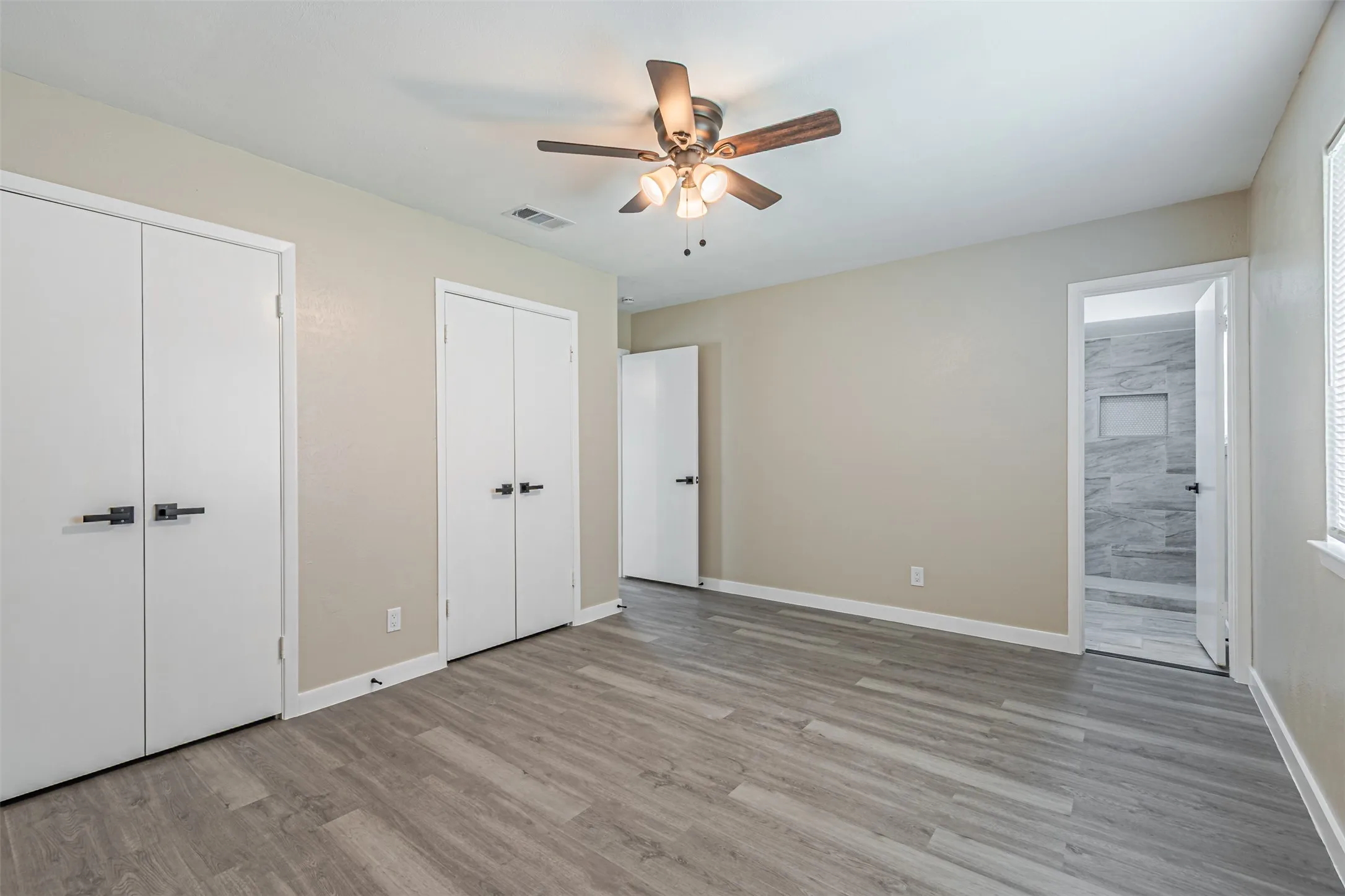 Room featuring wood-finish flooring, a ceiling fan with integrated lighting, dual closet doors with matte black hardware, and an entry to a tiled shower area
