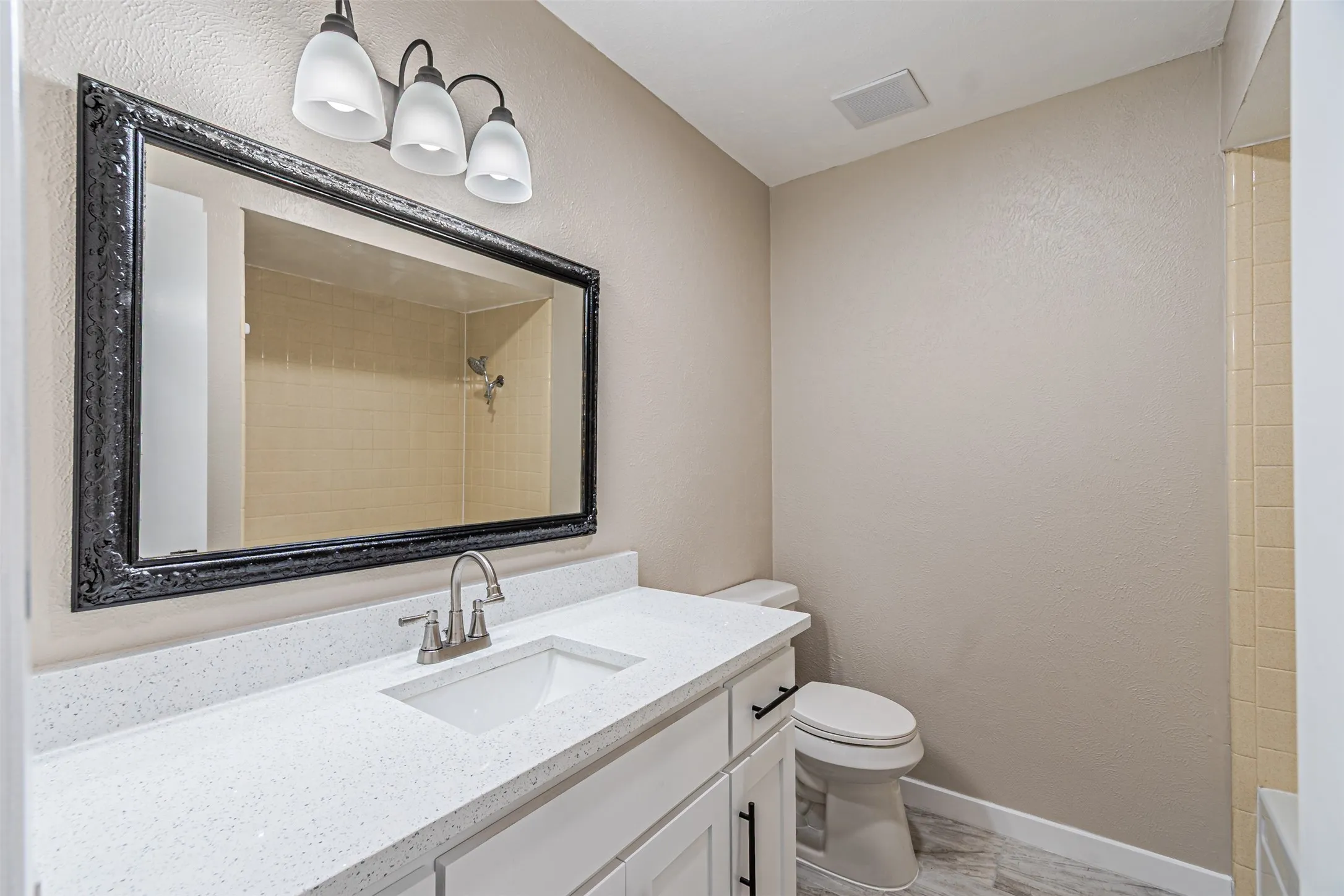Bathroom featuring a vanity with a white countertop and rectangular undermount sink, black-framed mirror, three-light fixture, and tile shower surround