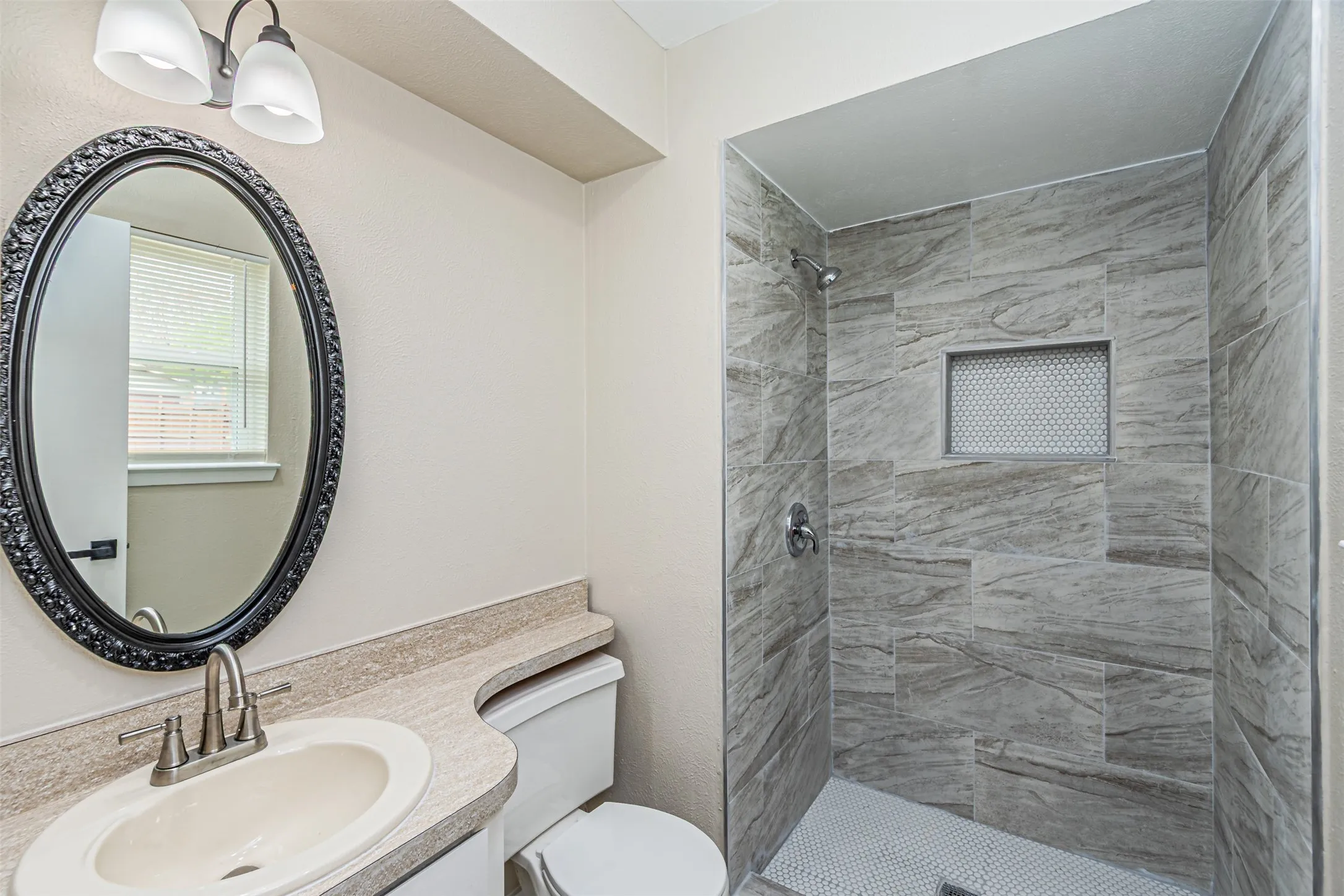 Bathroom featuring a walk-in shower with stone-patterned wall tile, a recessed niche, a neutral-toned countertop with an integrated basin, and an oval mirror with a decorative frame