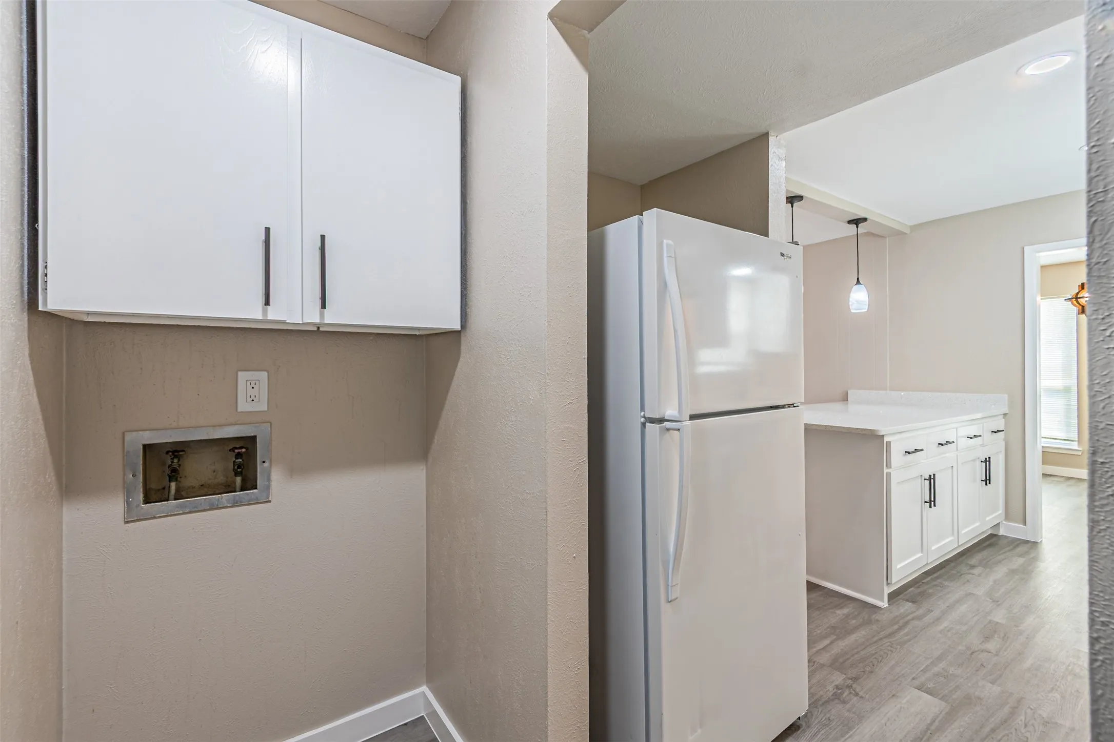 Dedicated utility area featuring white wall-mounted cabinetry and recessed plumbing connections, paired with a white refrigerator and kitchen cabinetry with light-toned countertops, finished with wood-finish flooring