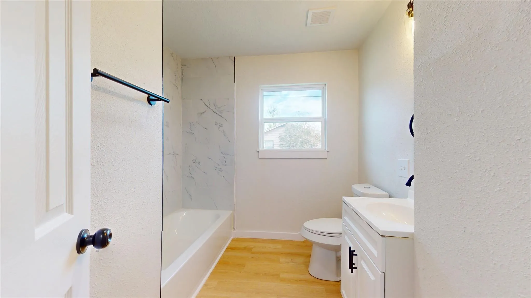 Bathroom featuring wood-finish flooring, a white vanity with an integrated countertop, matte black hardware, and a tub enclosure with light-toned patterned wall surfacing