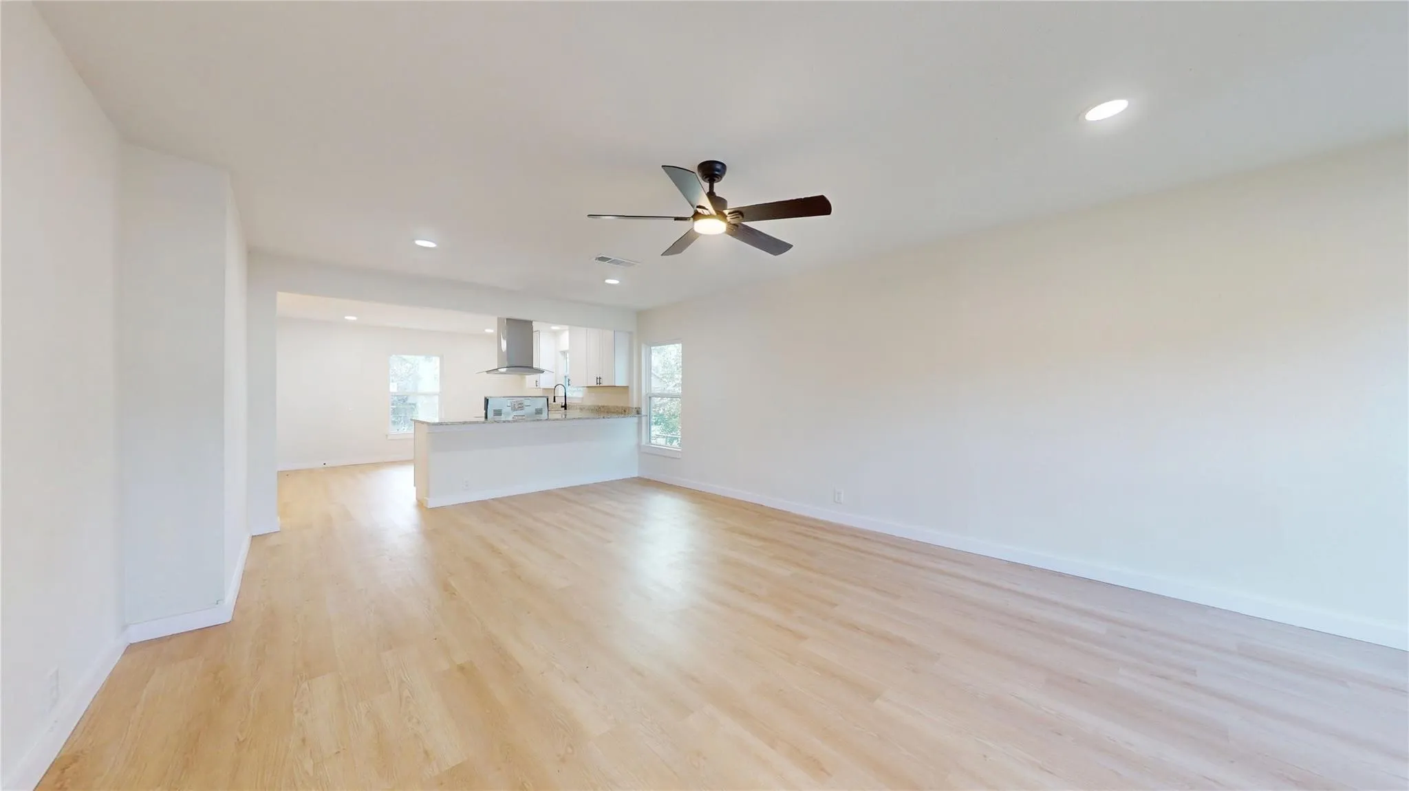 Open-concept area featuring light-tone wood-finish flooring, recessed lighting, a ceiling fan with integrated light, and a kitchen peninsula with cabinetry