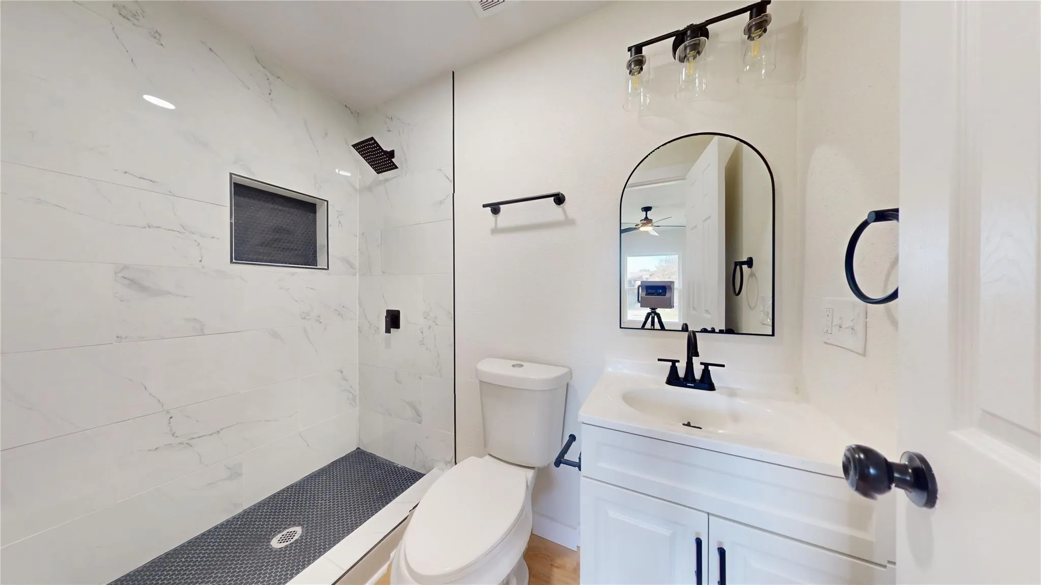 Walk-in shower with floor-to-ceiling stone-pattern tile, contrasting dark mosaic floor, built-in recessed wall niche, arched vanity mirror, and matte black hardware