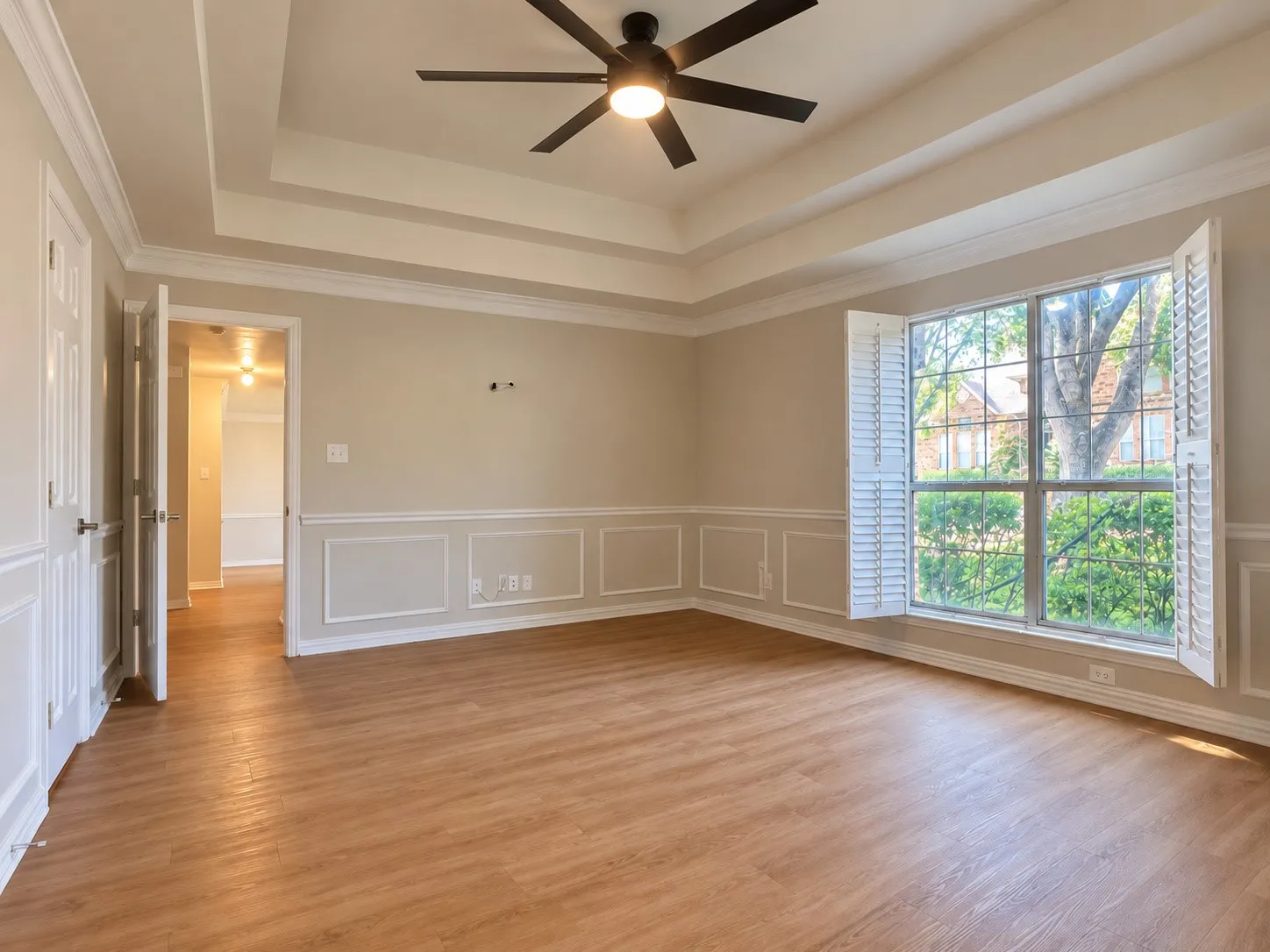 Tray ceiling, wood-finish flooring, wainscoting, multi-pane window with plantation shutters, and six-blade ceiling fan