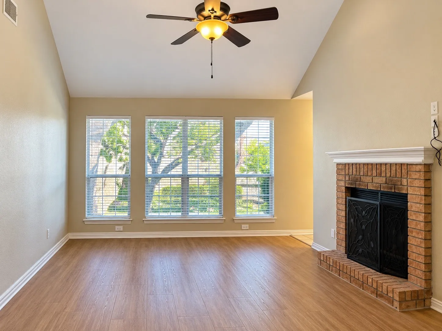 Vaulted ceiling featuring a central lighted fan, wood-finish flooring, triple window configuration, and brick fireplace with a white mantel