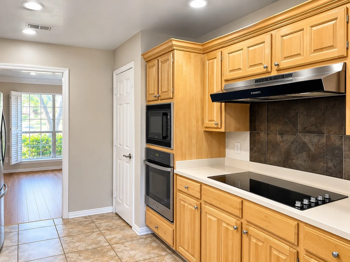 Kitchen featuring light-finish cabinetry, built-in wall oven and microwave, smooth cooktop with stainless steel range hood, stone tile backsplash, and tile flooring