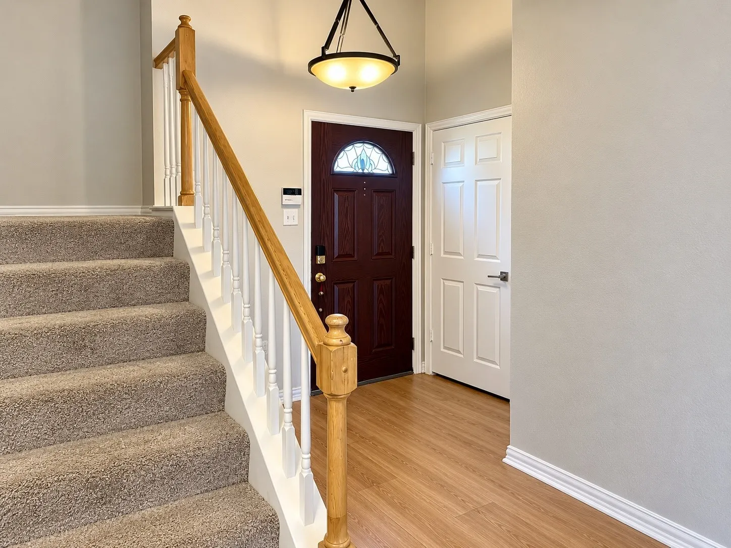 Entryway featuring wood-finish flooring, a wood-toned entry door with a decorative glass insert, a staircase with carpeted treads, a white balustrade with a wooden handrail, and a bowl-style pendant light fixture