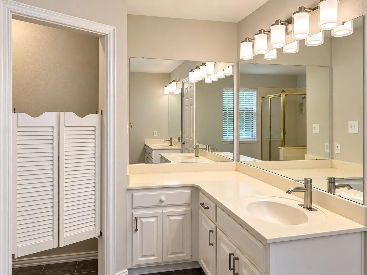 L-shaped vanity with integrated sinks, neutral solid-surface countertops, white cabinetry with metallic hardware, large wall-mounted mirrors, and multi-light vanity fixtures