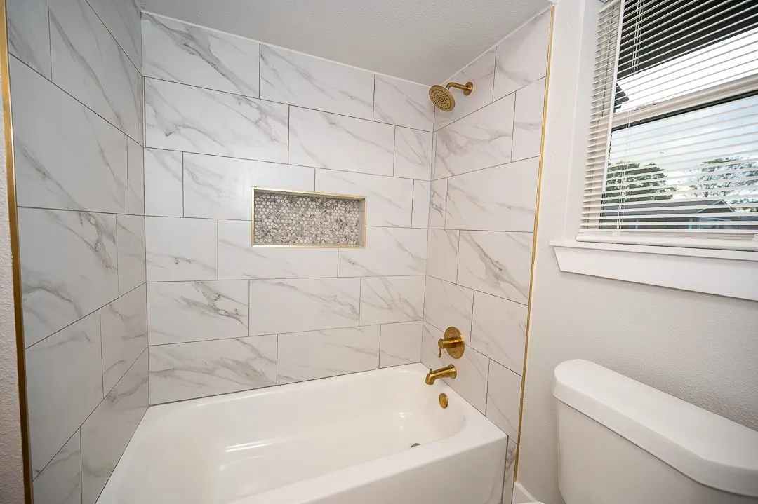 Bathtub enclosure featuring large-format stone-patterned wall tile, a recessed mosaic-tiled soap niche, and brass-finish plumbing fixtures