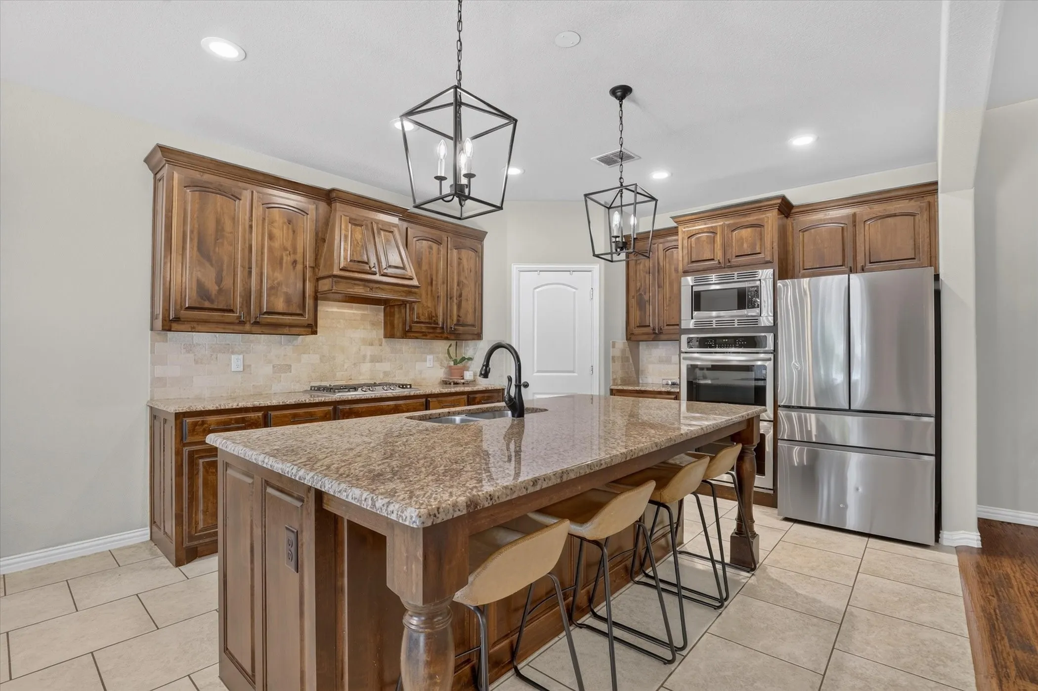 Kitchen featuring stained wood cabinetry, stone countertops, a center island with a dual-basin sink, built-in stainless steel appliances, and dual geometric pendant lighting