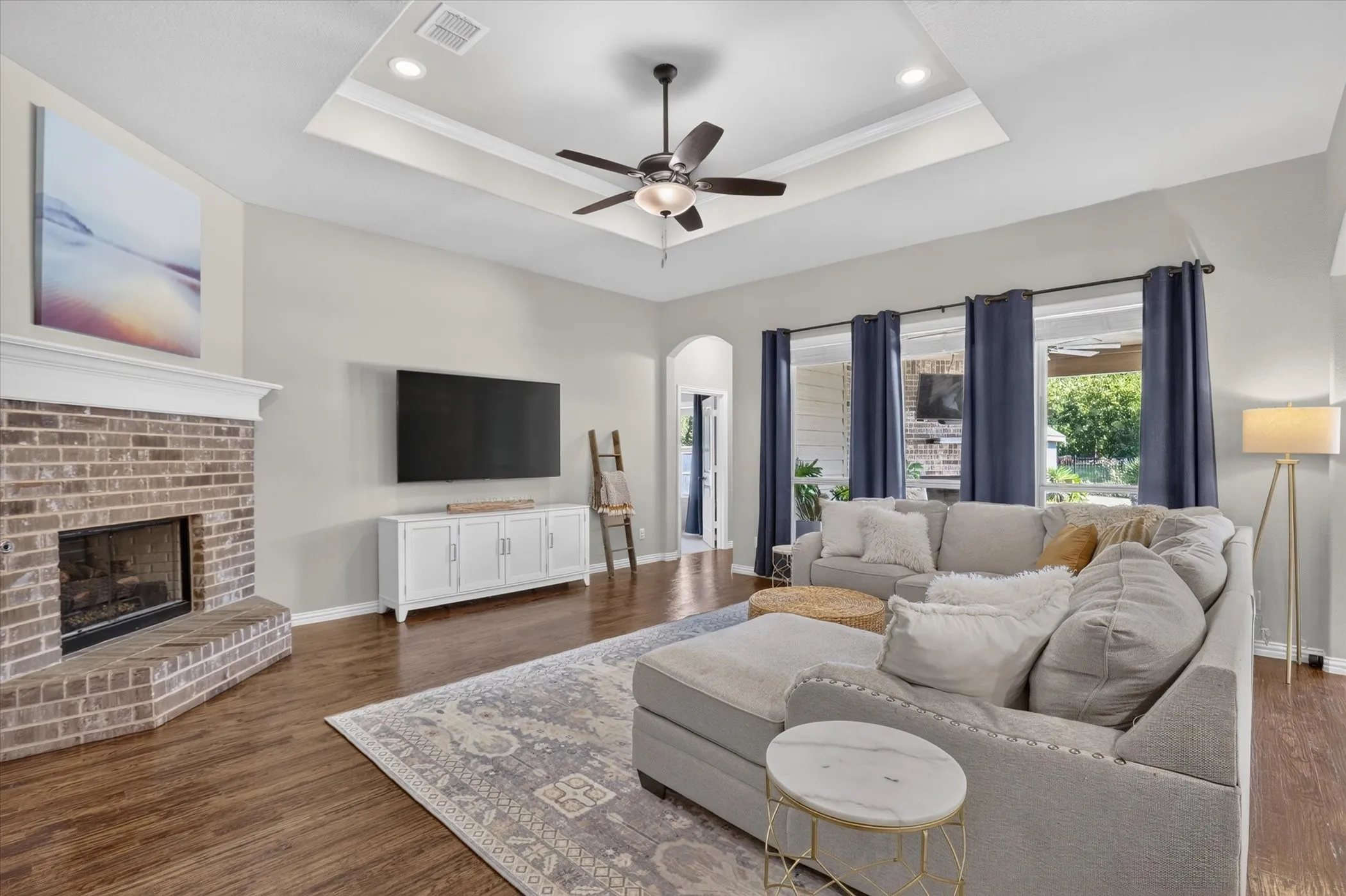 Living area featuring wood-finish flooring, a tray ceiling with a central fan, and a floor-to-ceiling brick fireplace with a mantel