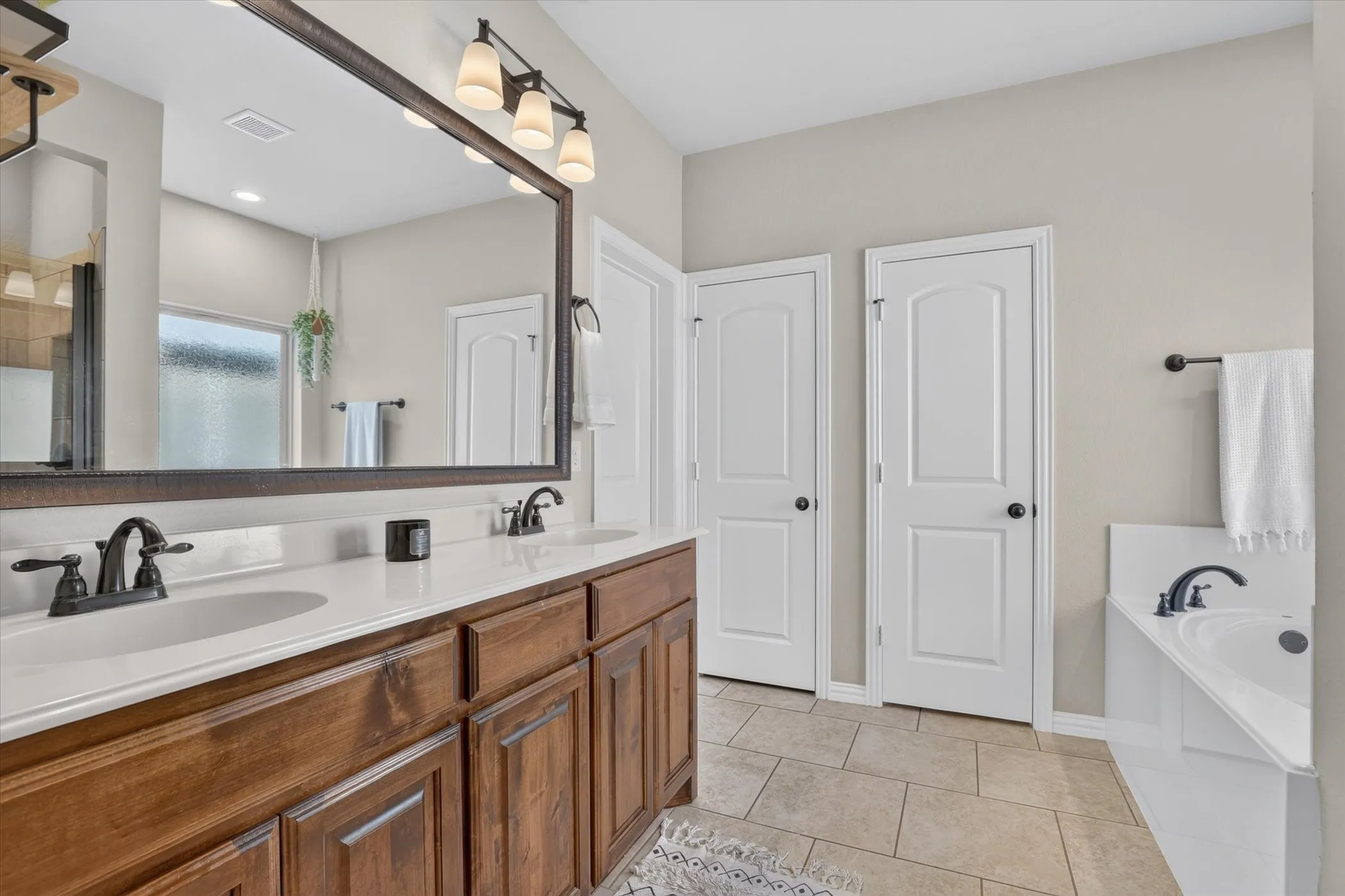 Bathroom featuring a dual-sink vanity with wood cabinetry and light-toned countertop, large framed wall mirror, three-light fixture, tile flooring, and a built-in soaking tub with deck-mounted faucet
