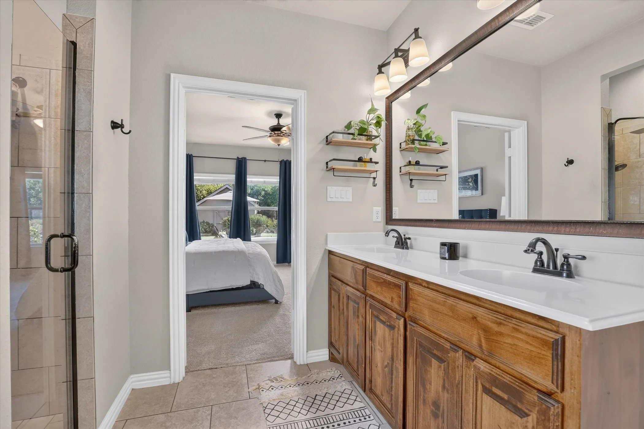 Dual-sink vanity with wood-finish cabinetry, solid surface countertops, oil-rubbed bronze fixtures, framed mirror, and walk-in shower with tiled surround