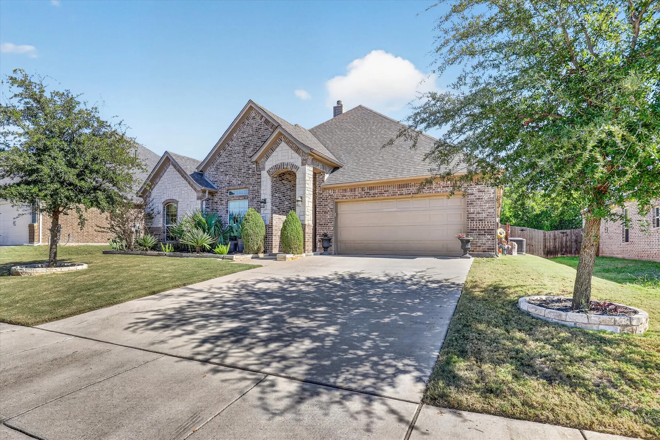 Brick and stone exterior featuring an arched entryway, shingle roof, concrete driveway, and landscaped front yard with stone-bordered tree wells