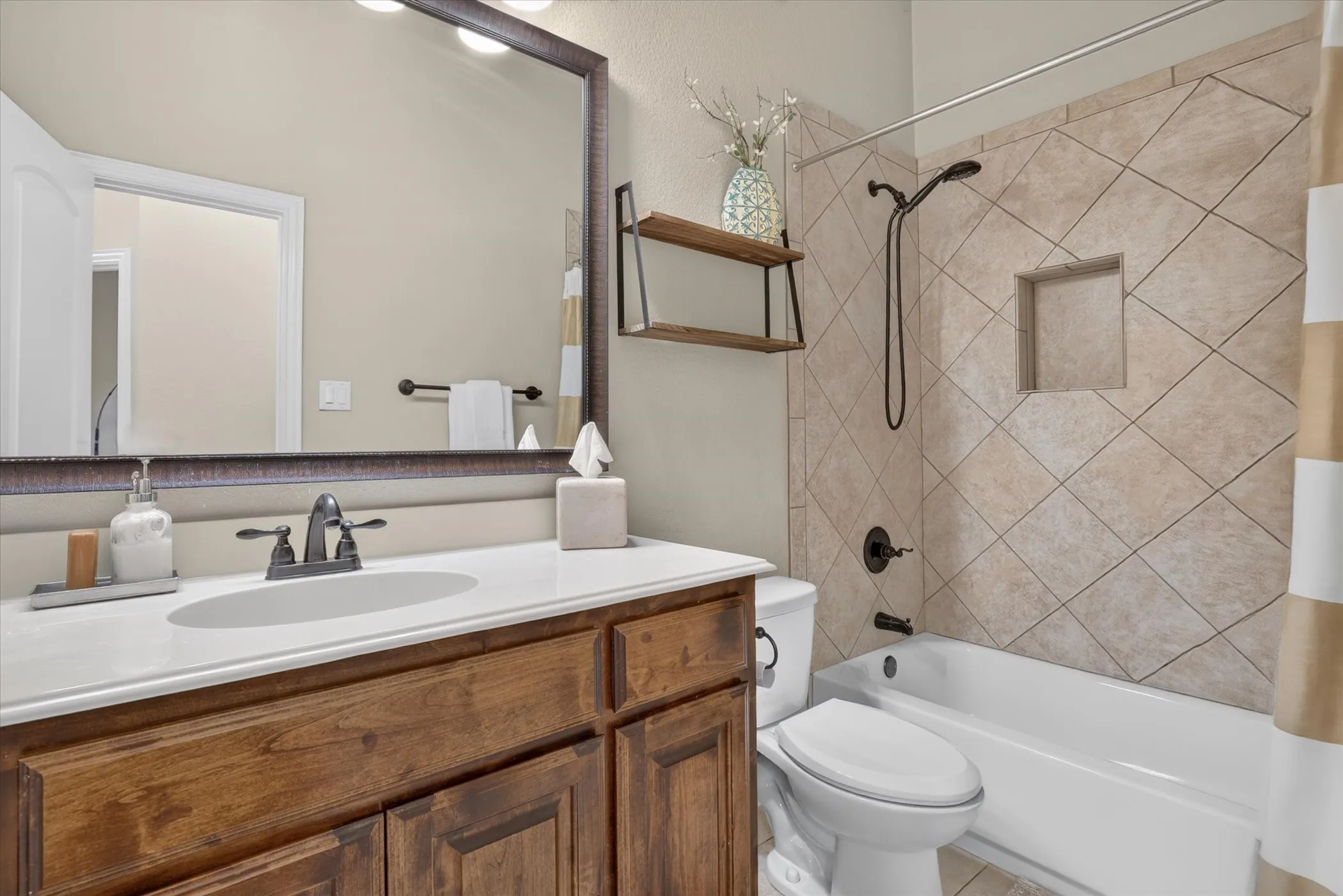 Bathroom featuring a wood-finish vanity with a stone countertop, an oval undermount sink, a large framed mirror, and a shower-tub combination with diamond-patterned wall tile and a built-in recessed niche