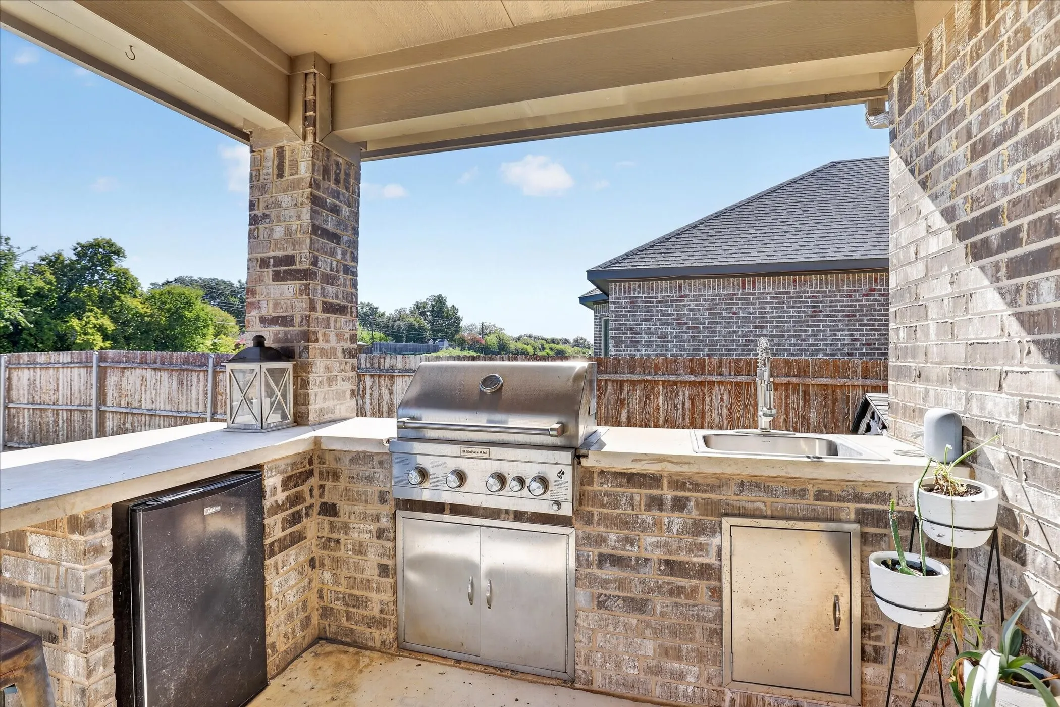 Covered outdoor kitchen featuring brick construction, a stainless steel gas grill, under-counter refrigeration, a built-in sink, and light-toned stone countertops
